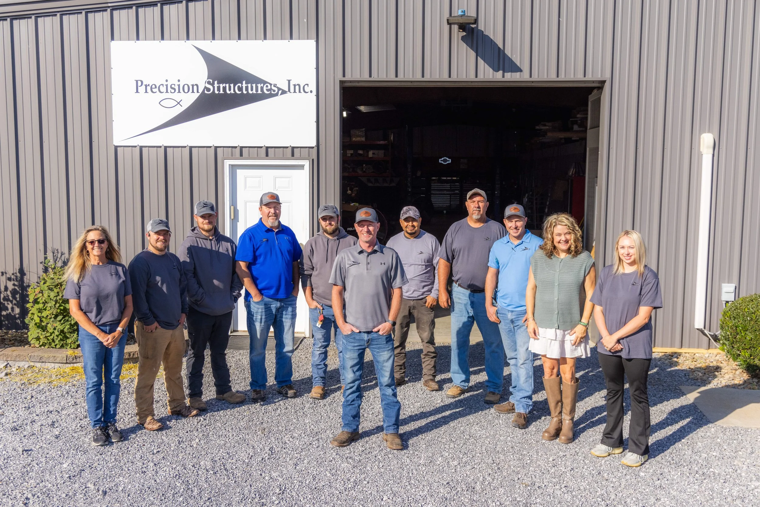A group of eleven people standing in front of a gray industrial building with a sign that reads 'Precision Structures Inc.' inside a parking area with gravel ground. The group includes men and women smiling and posing for the photo.