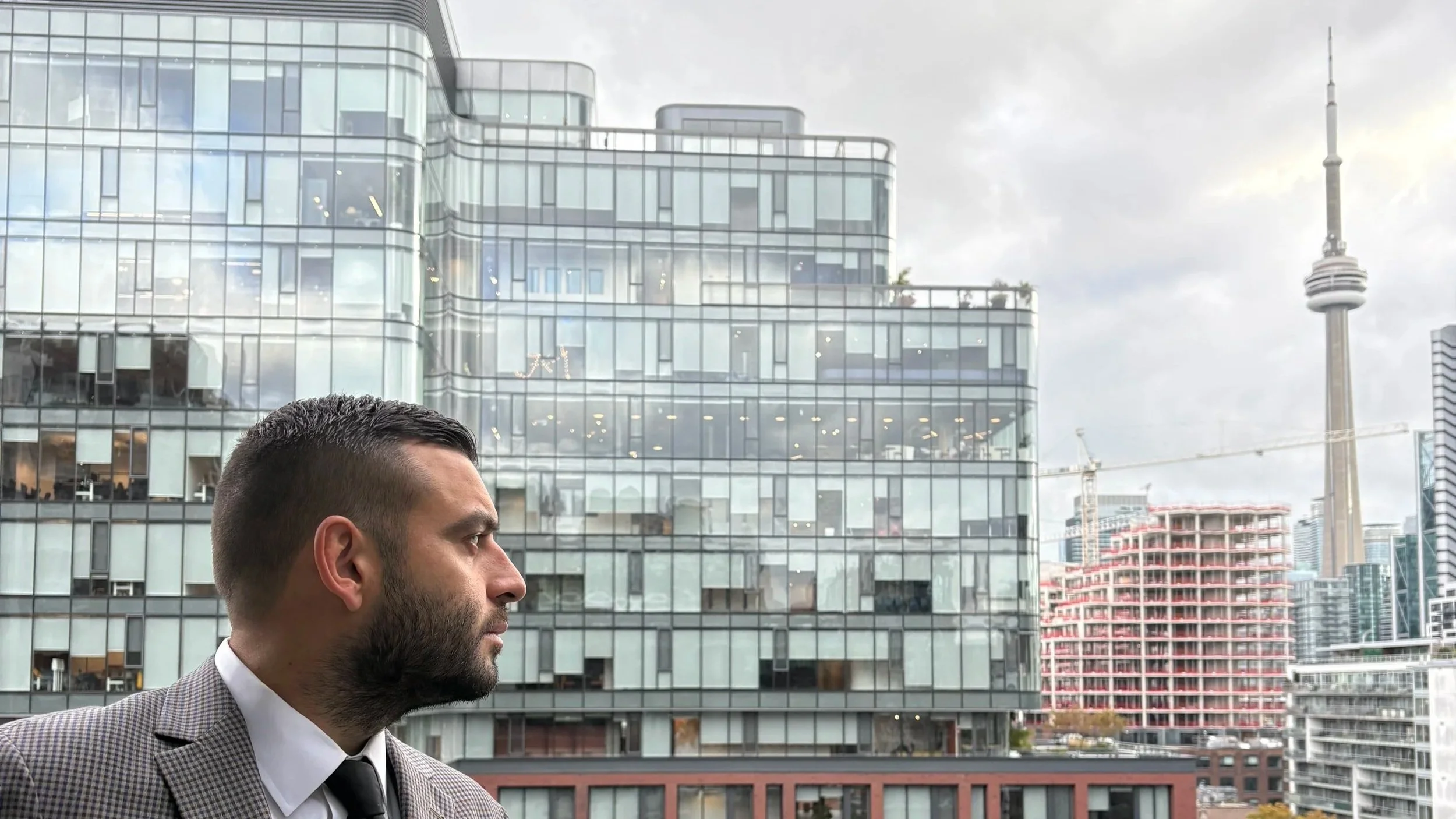 Profile of a man with a beard and short dark hair wearing a checkered blazer and black tie, standing against a cityscape background with modern glass buildings and a tall TV tower.