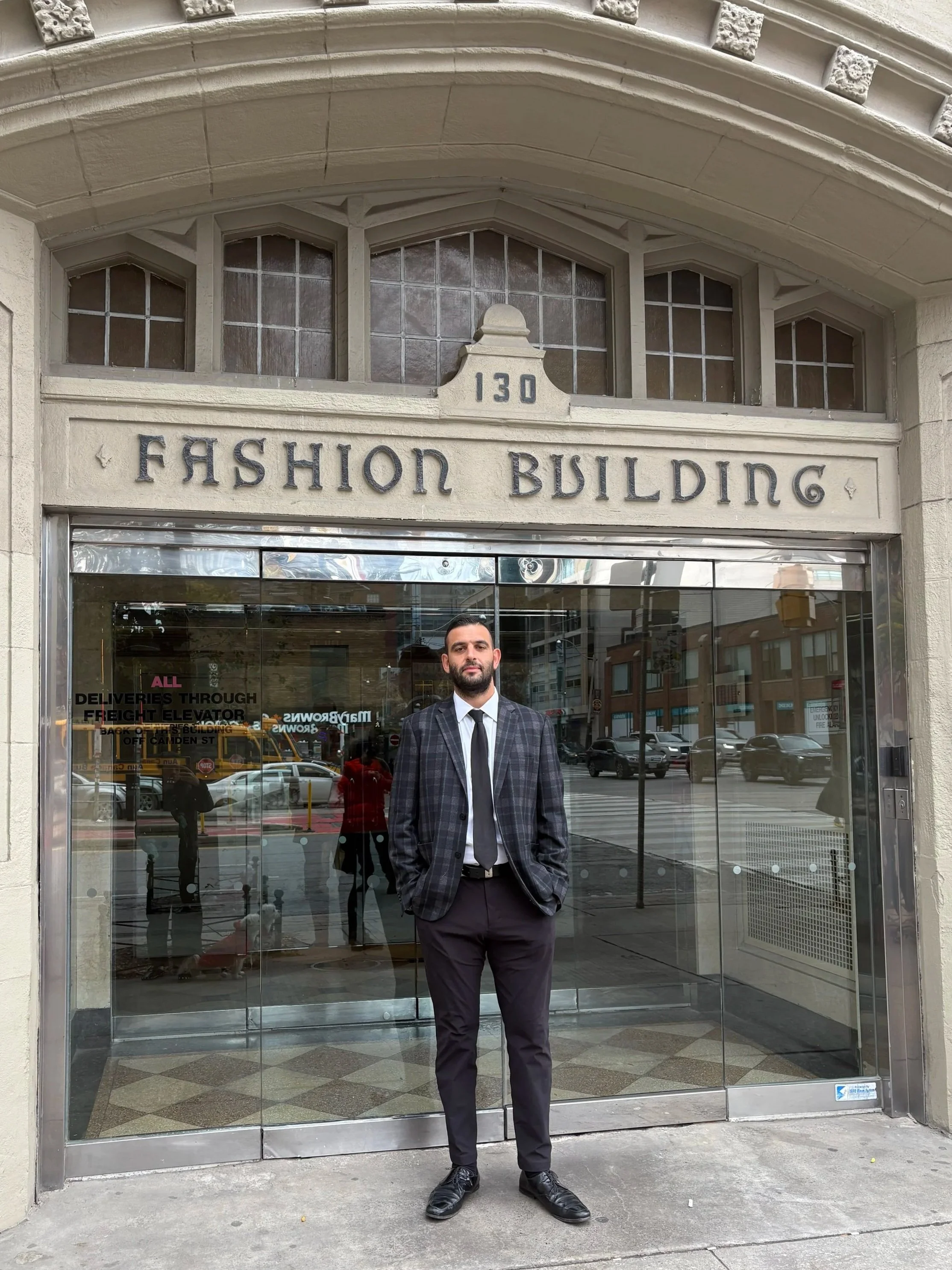 A man in a checkered suit and black tie standing in front of a glass entrance to the Fashion Building at 130. The building has a stone facade and the street reflection is visible on the glass.