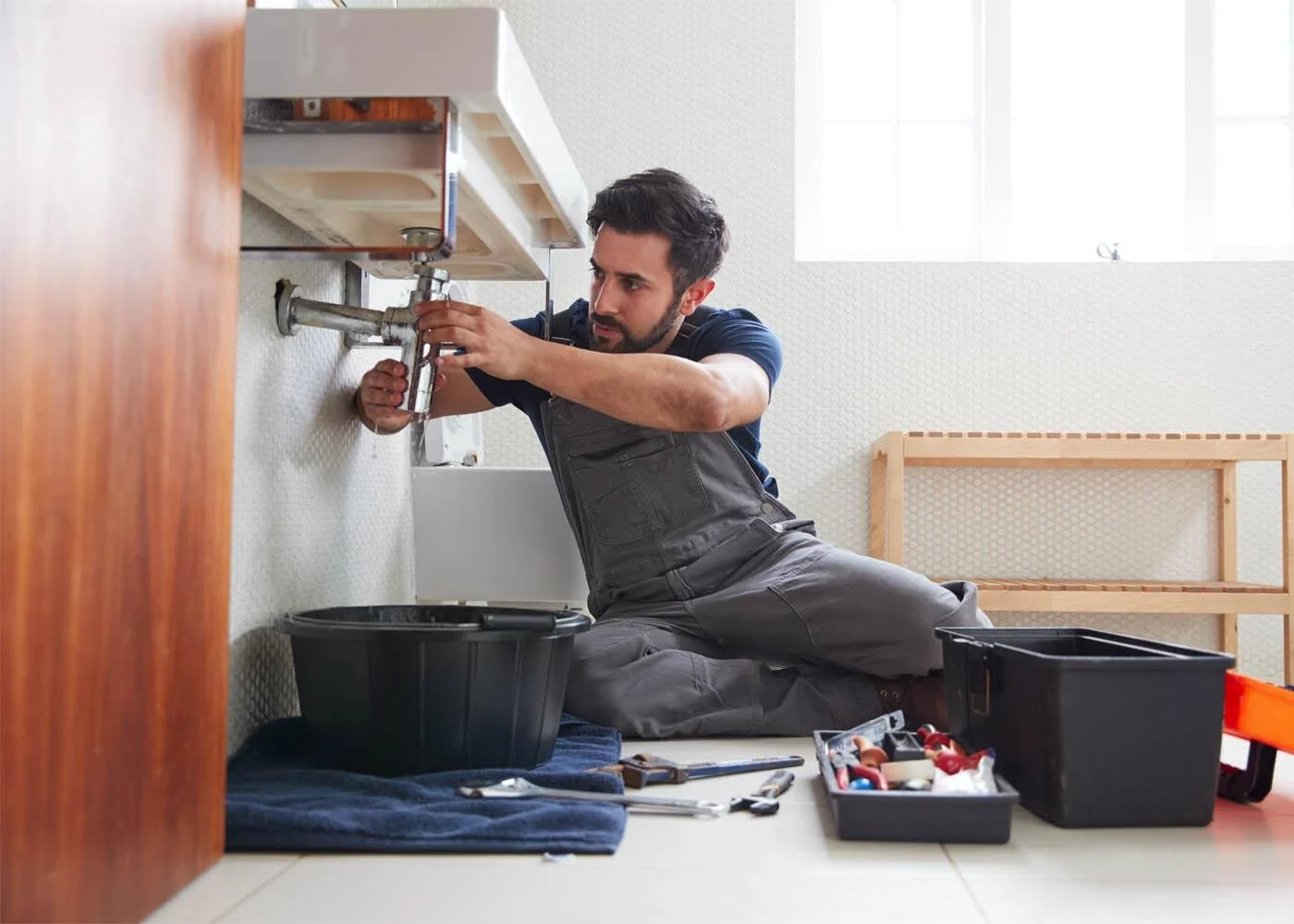 Man kneeling on the floor installing a kitchen sink under a cabinet, surrounded by tools and toolboxes.
