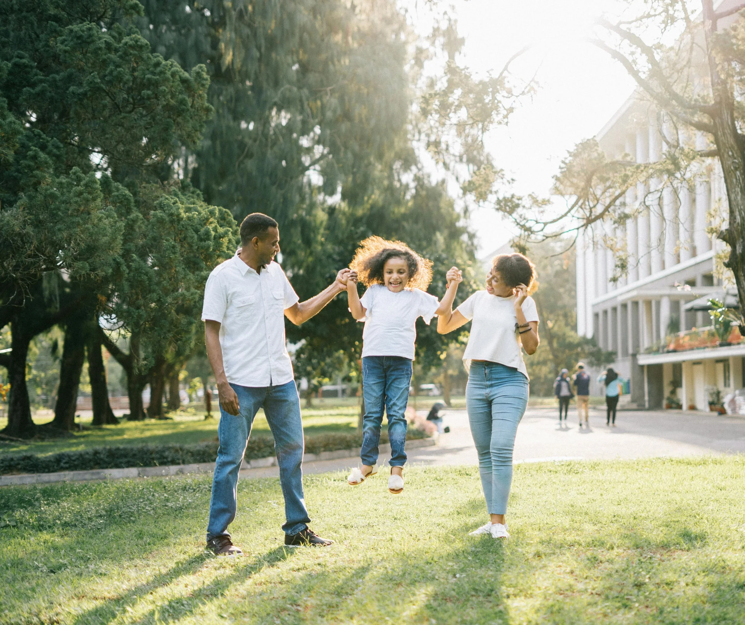 A family of three playing outdoors on a sunny day, with the father and mother holding their daughter by the hands and swinging her in the air on a grassy area.