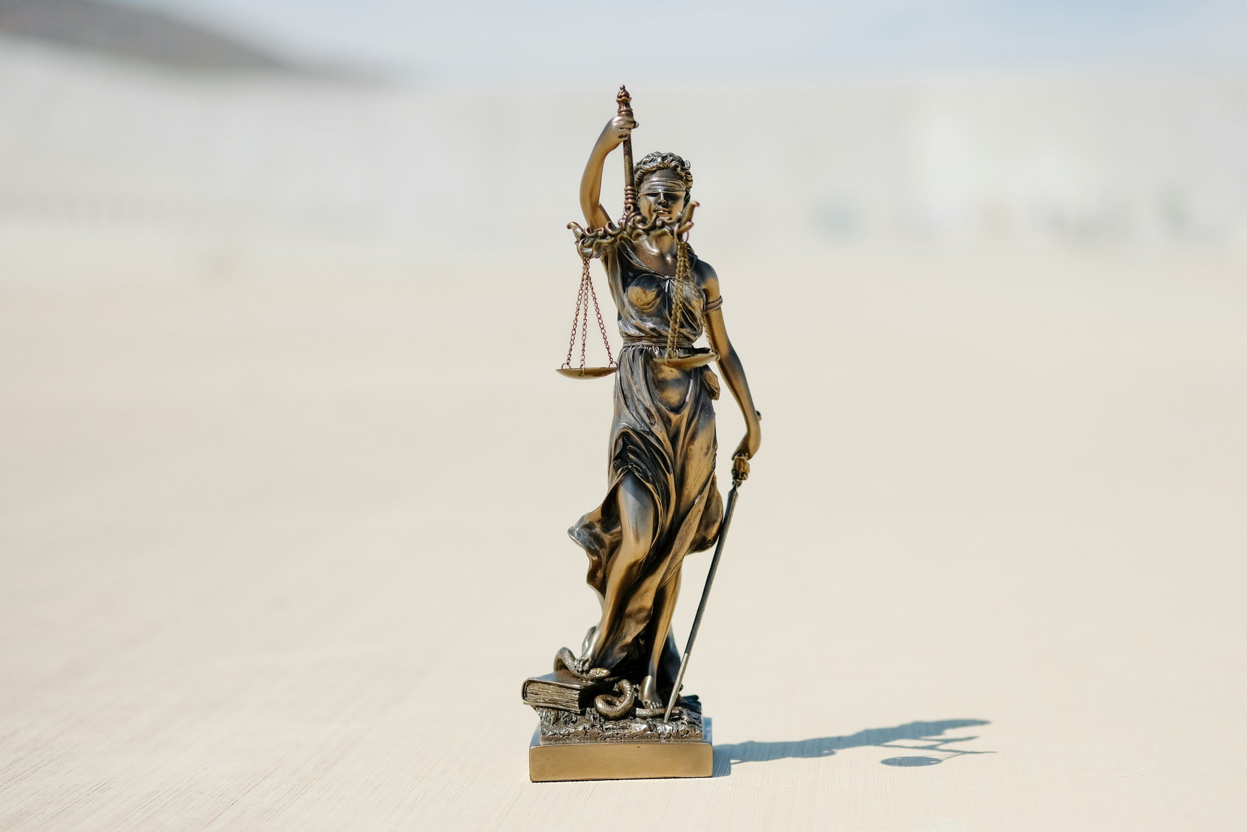 A small statue of Lady Justice holding a sword and a scale, standing on a book, casting a shadow on a light-colored surface.