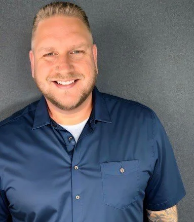 A man with short blond hair and a beard smiling, wearing a dark blue button-up shirt, standing against a gray background.