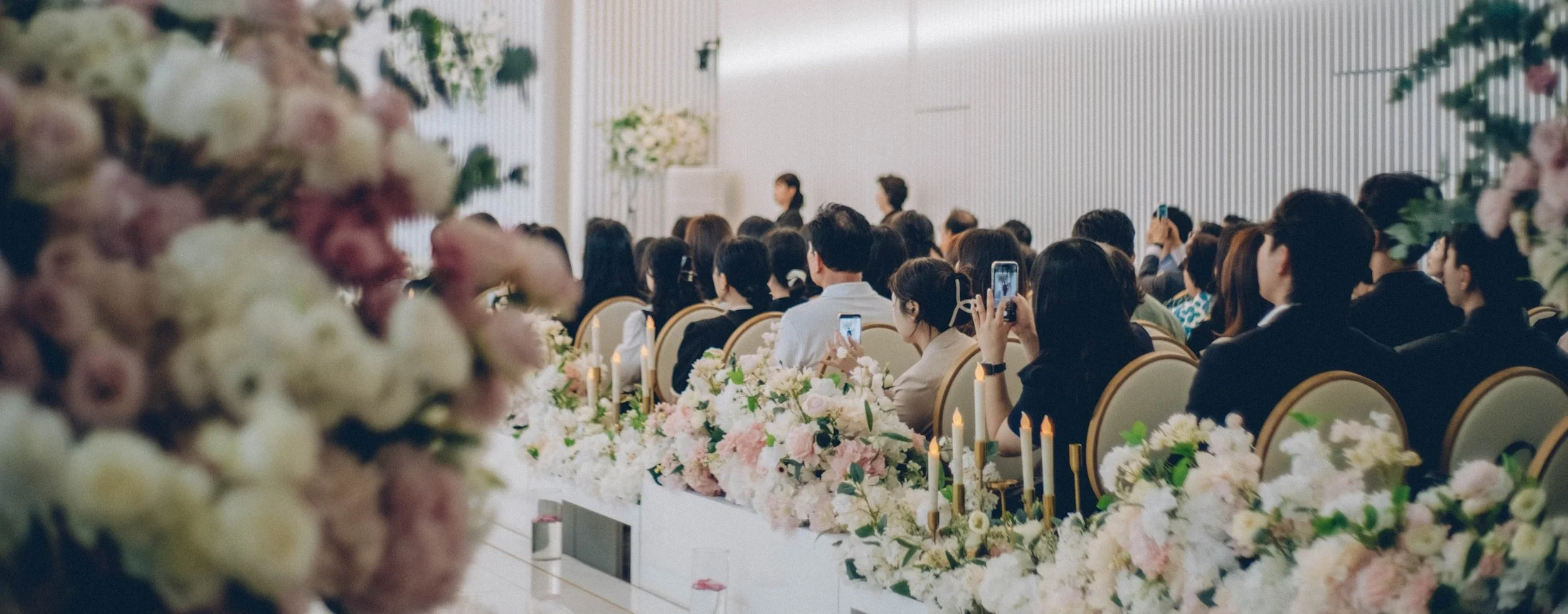 Crowd of people seated at wedding ceremony, some taking photos, decorated with flowers and candles.