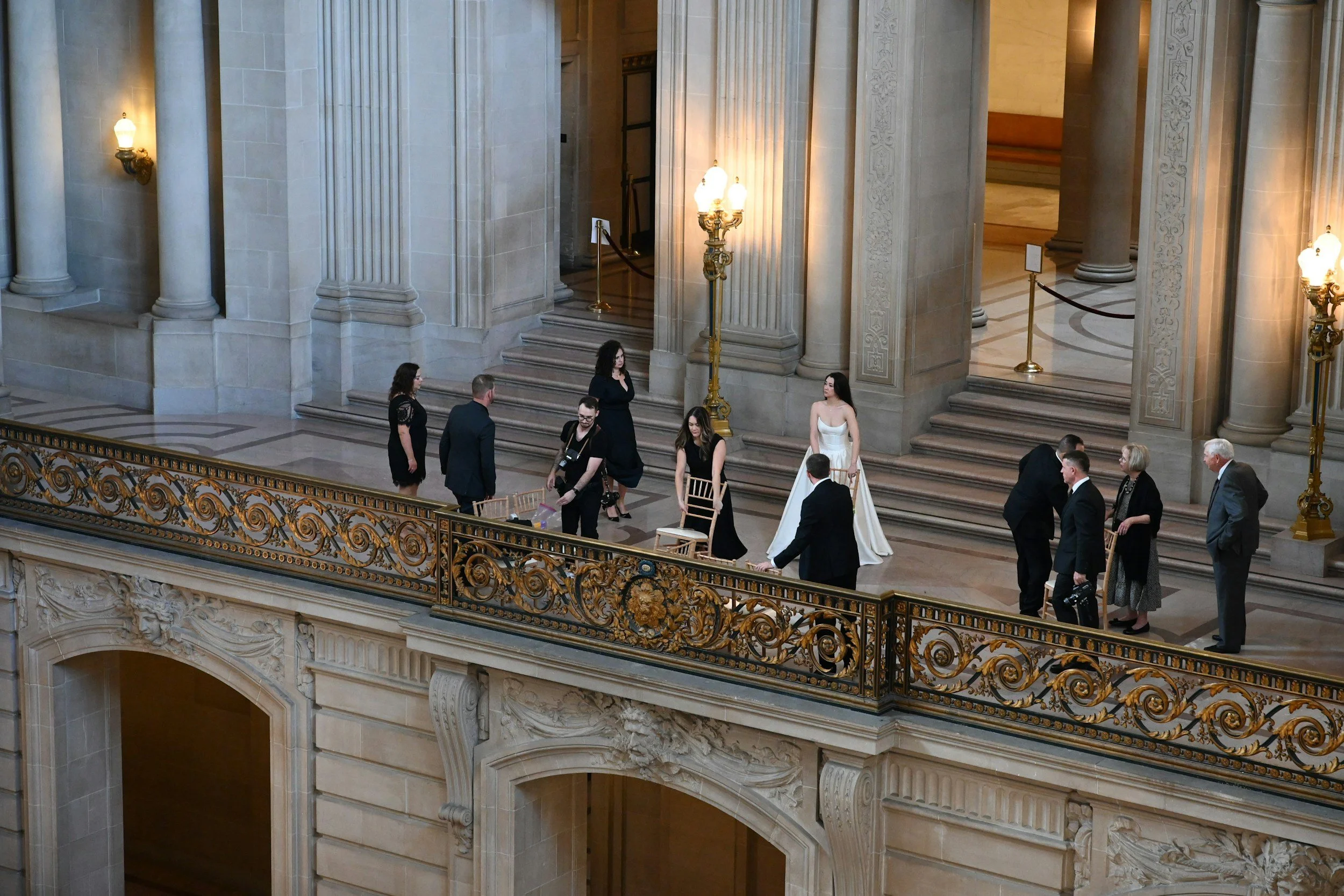 People dressed in formal attire inside a grand historic building with ornate railings, marble columns, and staircase.