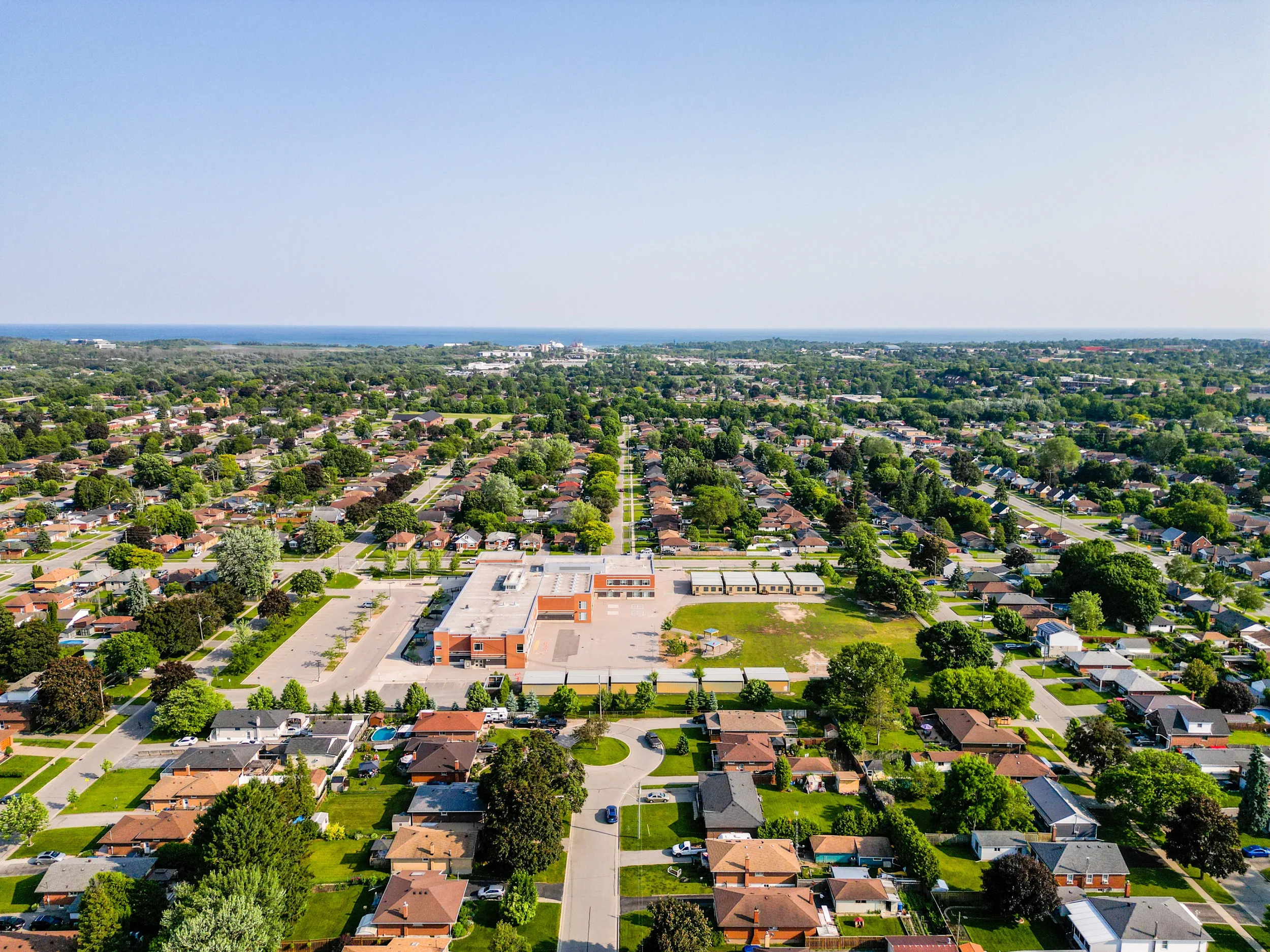 An aerial view of a suburban neighborhood with houses, trees, and a school in the center, near the coast.