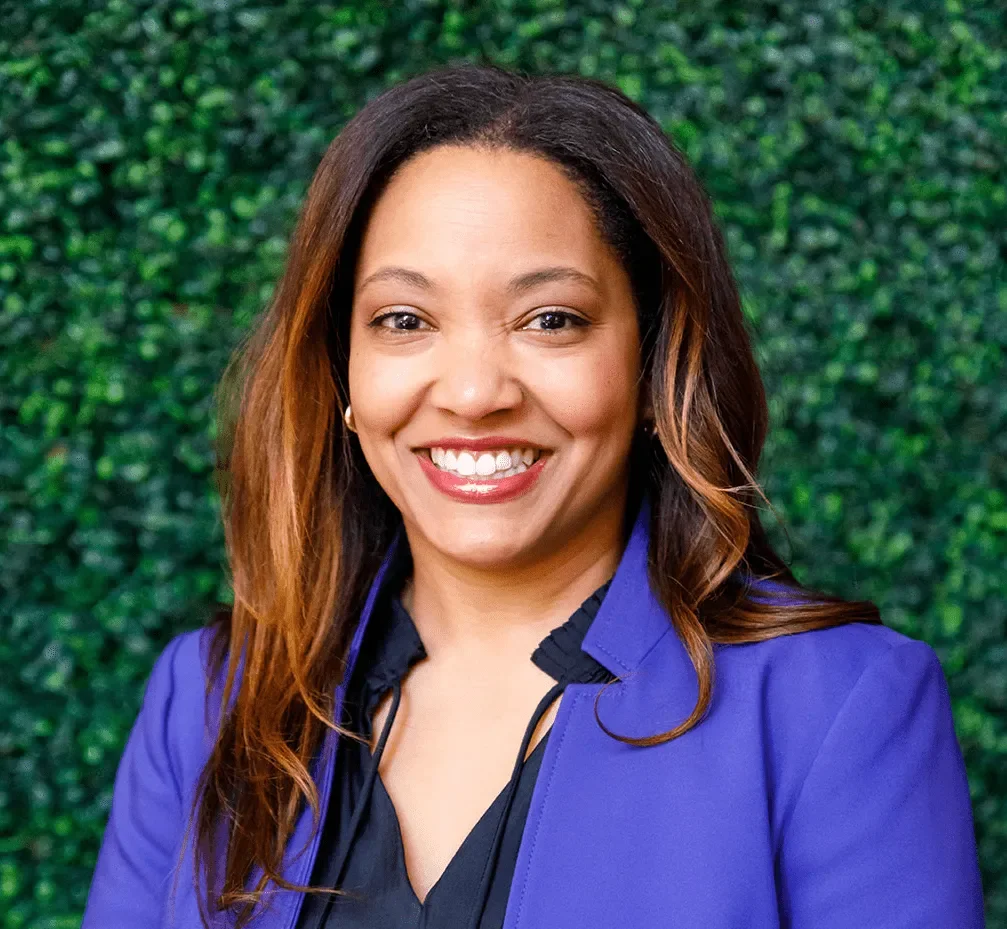A woman with shoulder-length brown hair smiling, wearing a dark top and a purple blazer. The background consists of green leafy foliage.