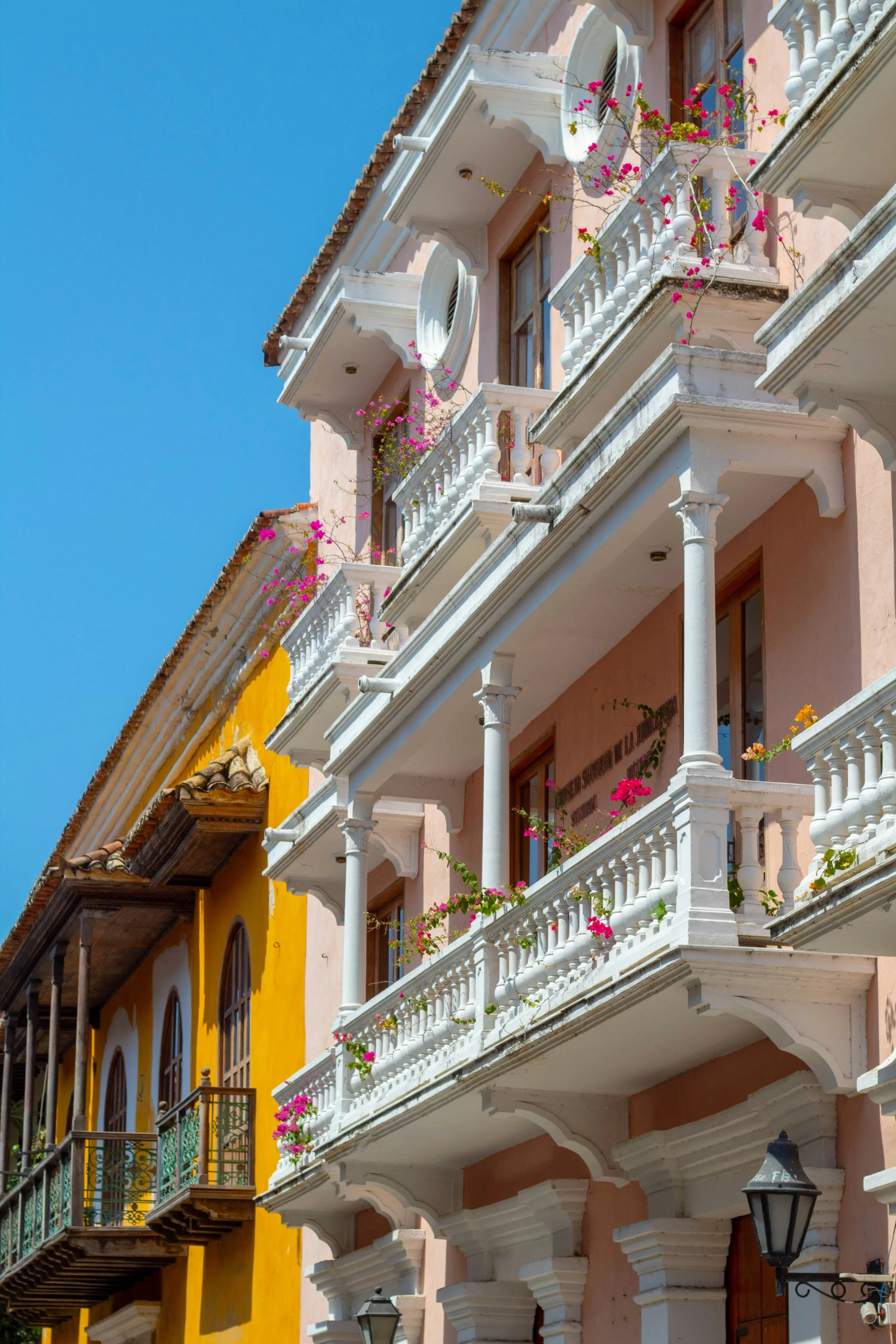 Colorful buildings with balconies, some adorned with flower pots and pink flowers, under a clear blue sky.