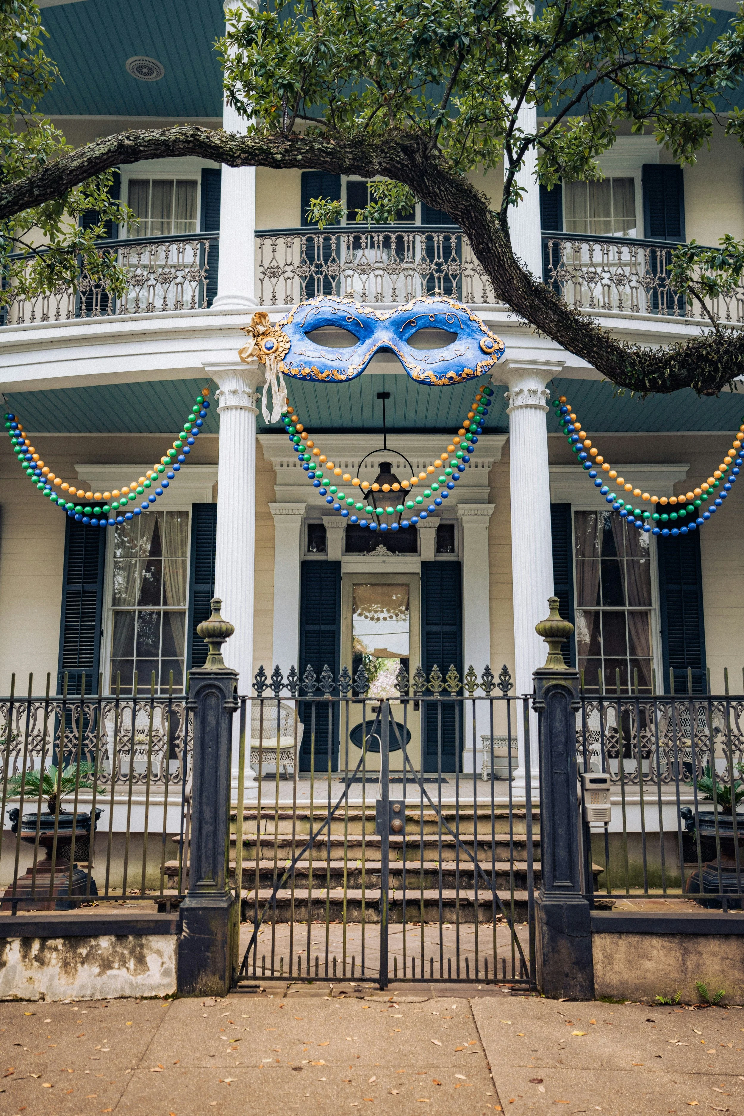 A decorated house with Mardi Gras beads draped on the porch and a Mardi Gras mask hanging in the center, celebrating Carnival.