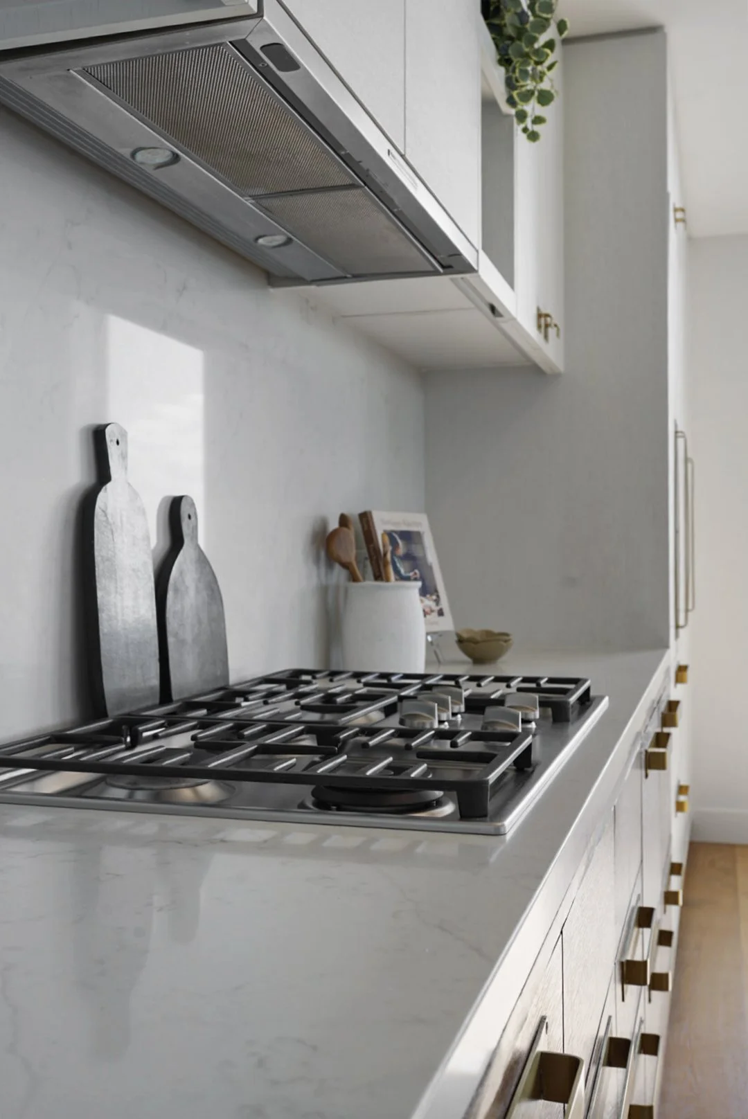 Modern kitchen countertop with a gas stove, two metal cutting boards, and a white container holding cooking utensils, with a plant and a cookbook in the background.