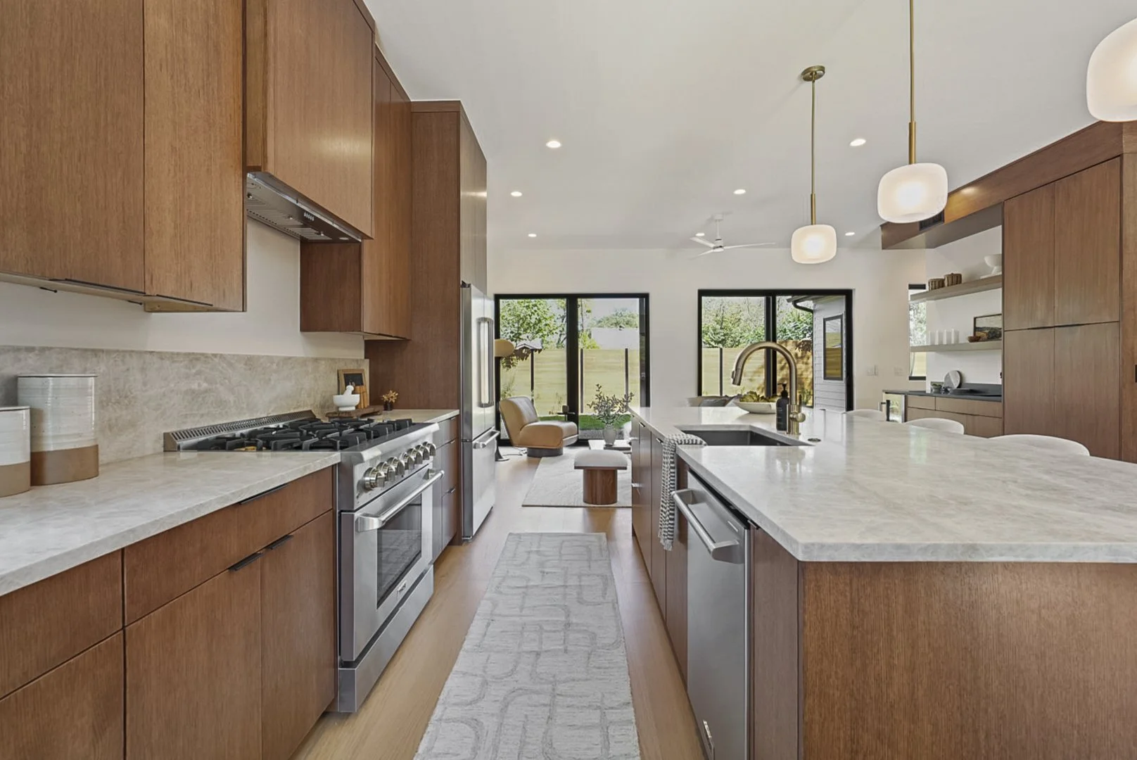 Modern kitchen with wooden cabinets, stainless steel appliances, and a marble kitchen island with a sink. There are pendant lights, and in the background, windows and a small seating area with a chair and a view of a backyard.