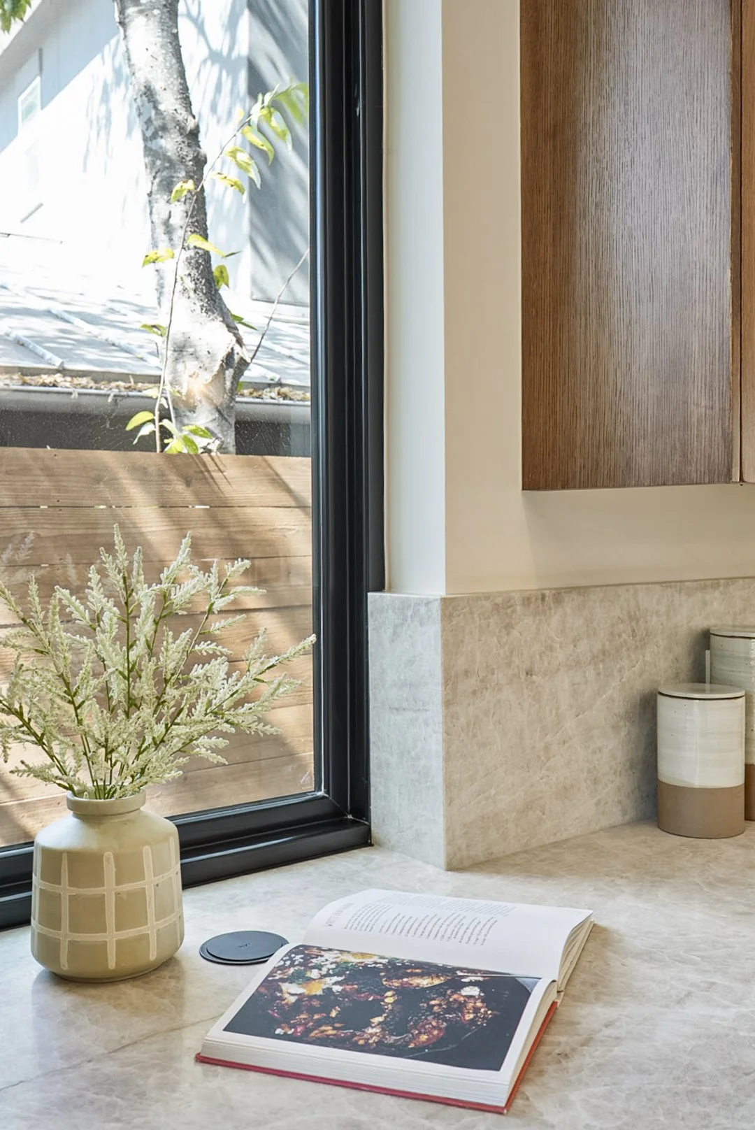A windowsill with a beige vase holding white and green plant, an open book showing a colorful image, a small round black object, and two white and beige container jars, with a view of a wooden fence and tree outside.