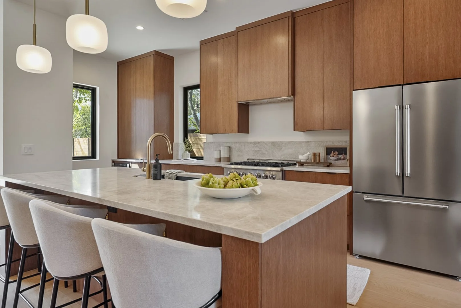 Modern kitchen with wooden cabinets, a large granite island with seating, stainless steel refrigerator, and black-framed windows with outdoor greenery.