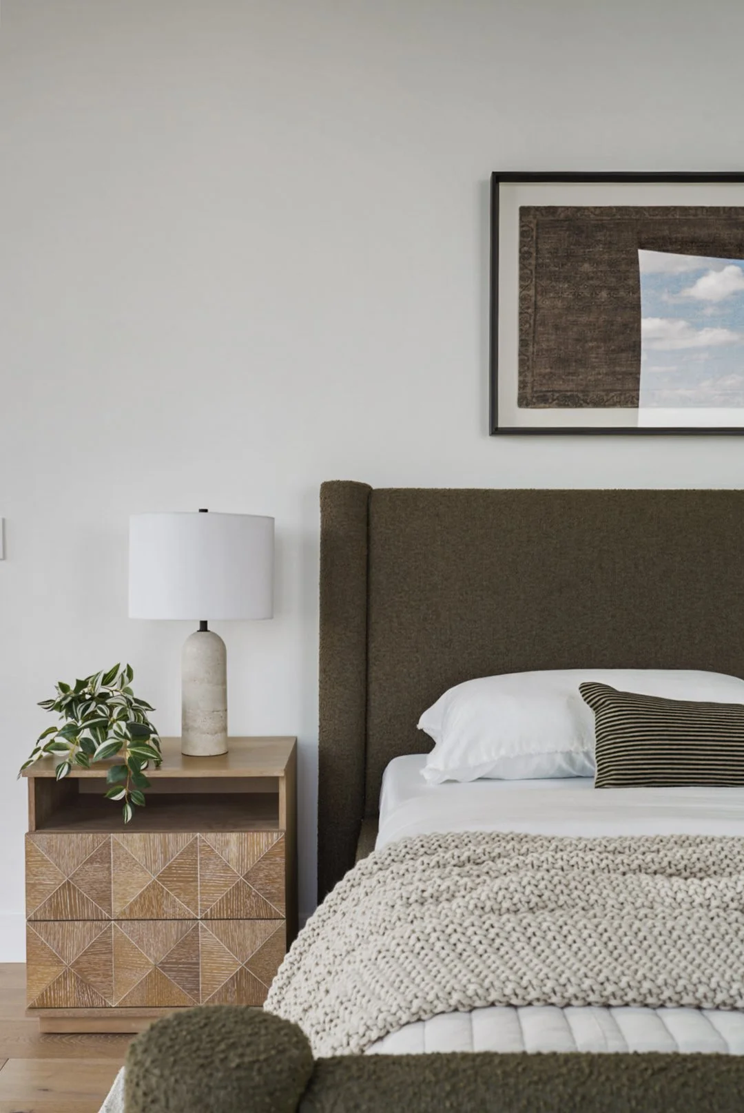 A bedroom with a brown upholstered headboard, white bedding, a beige chunky knit blanket, a black and beige striped pillow, a wooden nightstand with a textured drawer, a white lamp, a potted plant, and a framed print of a landscape with sky and cloud