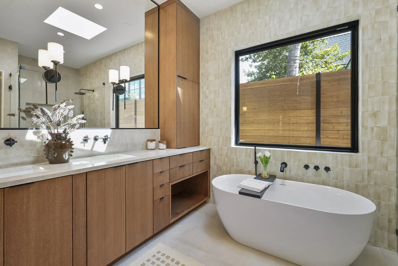 Modern bathroom featuring a freestanding bathtub, wooden cabinetry, a large window with a wooden privacy screen, and a marble countertop with a vase of flowers.