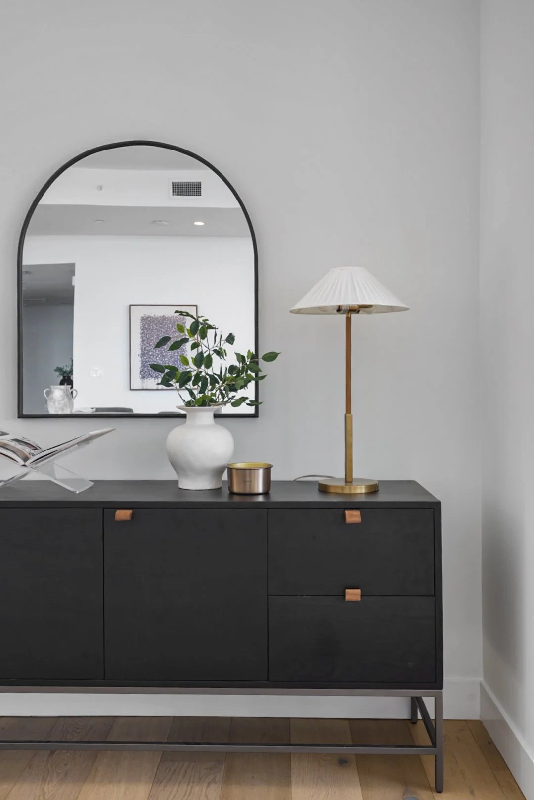 A modern black sideboard with wooden accents on the drawer handles, placed against a light gray wall. On top, there is a white vase with green leafy branches, a small metallic container, and an open book on a stand. A tall brass and white table lamp 
