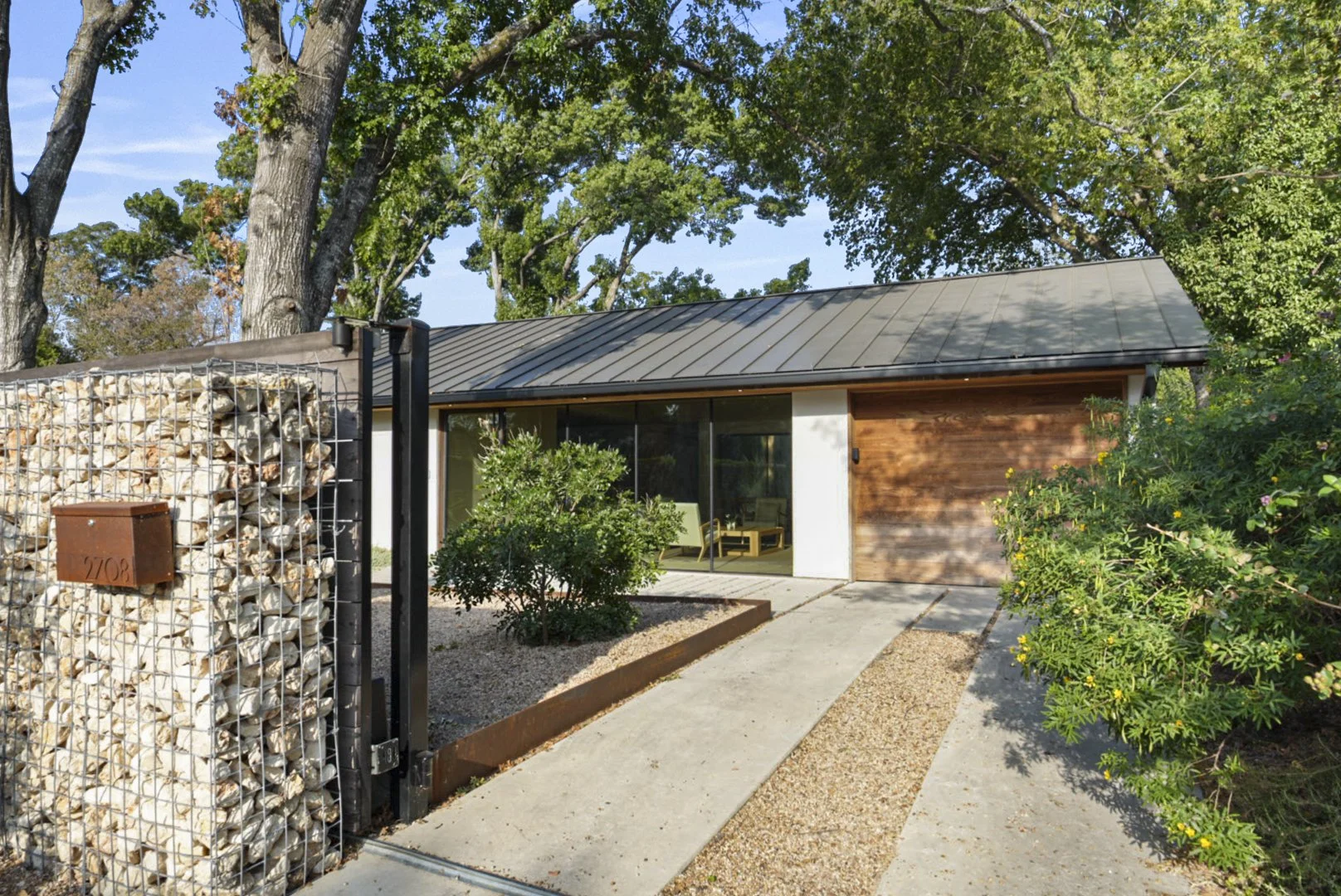Modern single-story house with large glass window, wooden siding, and metal roof, surrounded by trees and greenery.