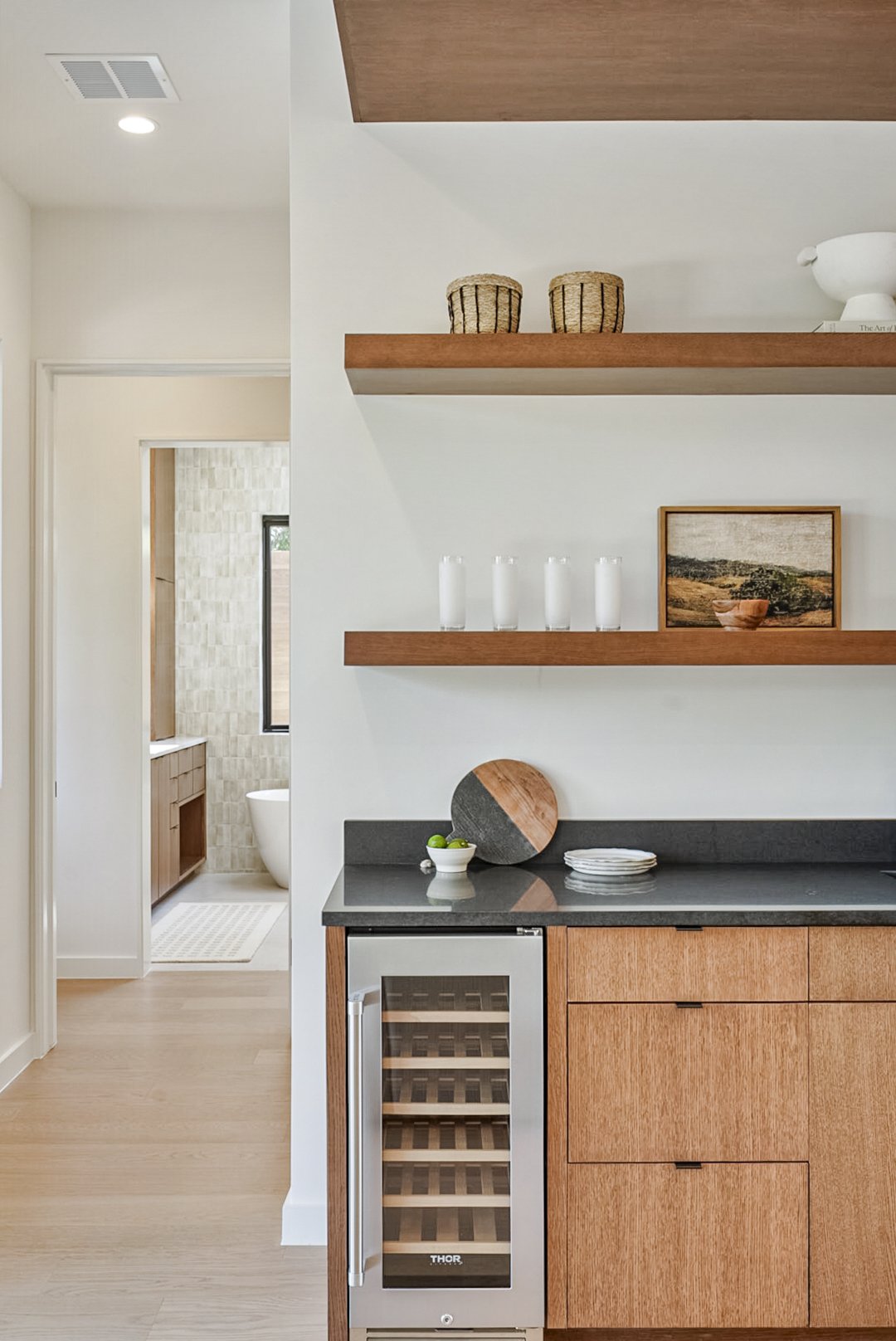 A kitchen corner with wooden cabinets, a wine cooler, and floating wooden shelves with decorative items.