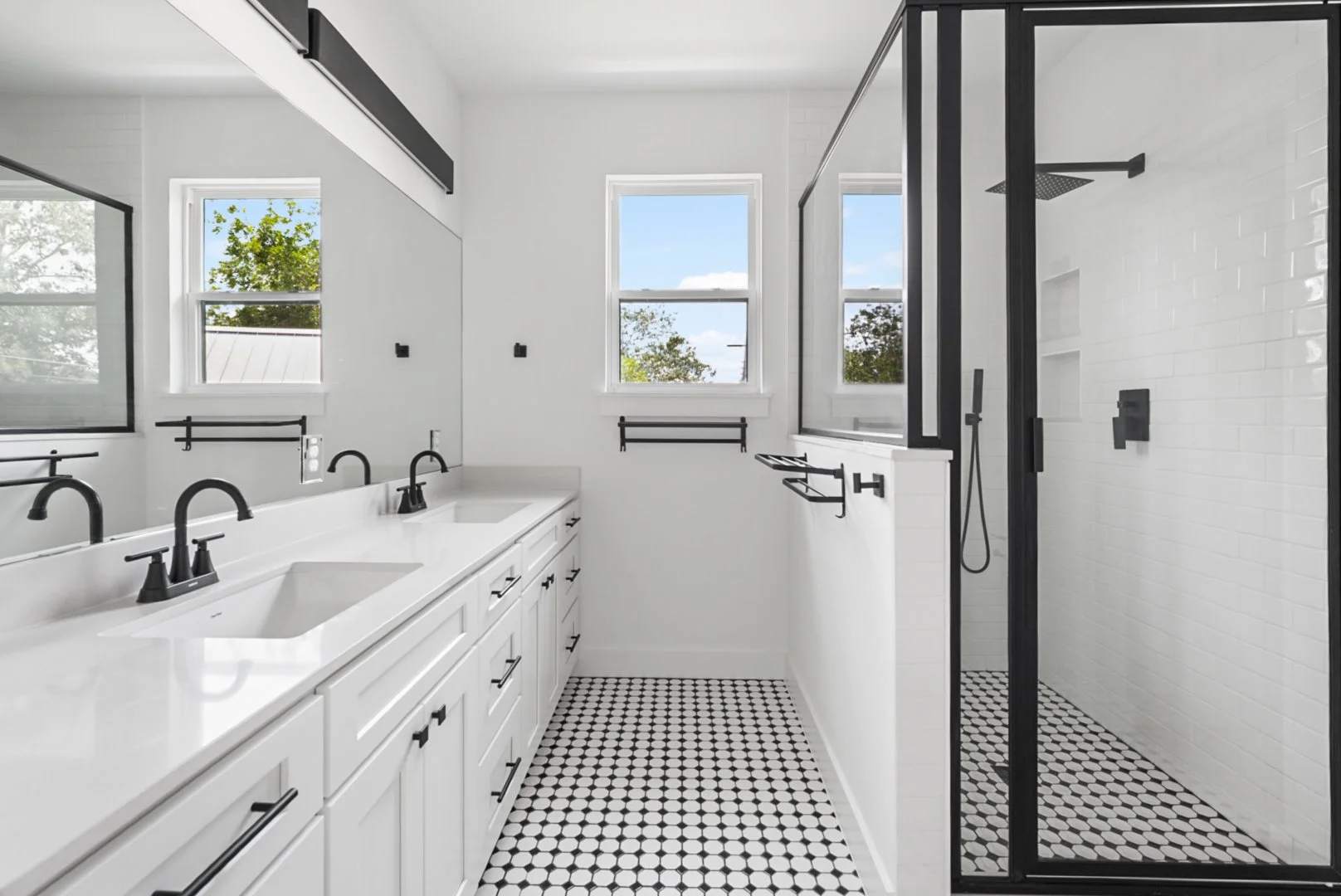 Modern bathroom with white cabinets, black fixtures, and black and white tiled floor. Features a double sink vanity, a walk-in shower with a glass door, and three windows with views of trees and a blue sky.