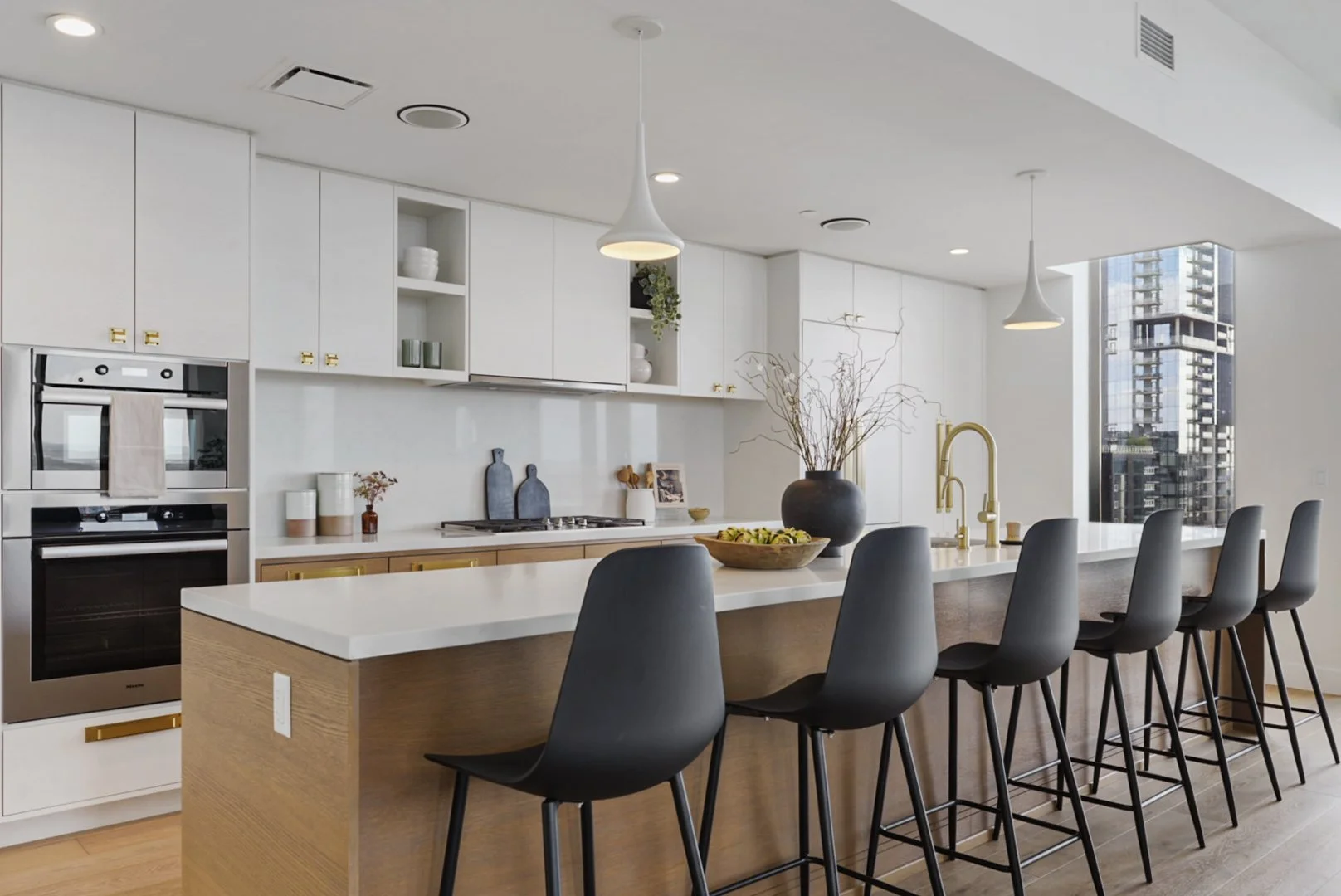 Modern kitchen with white cabinets, a large island with a wooden base, black chairs, a gold faucet, and city view through large windows.