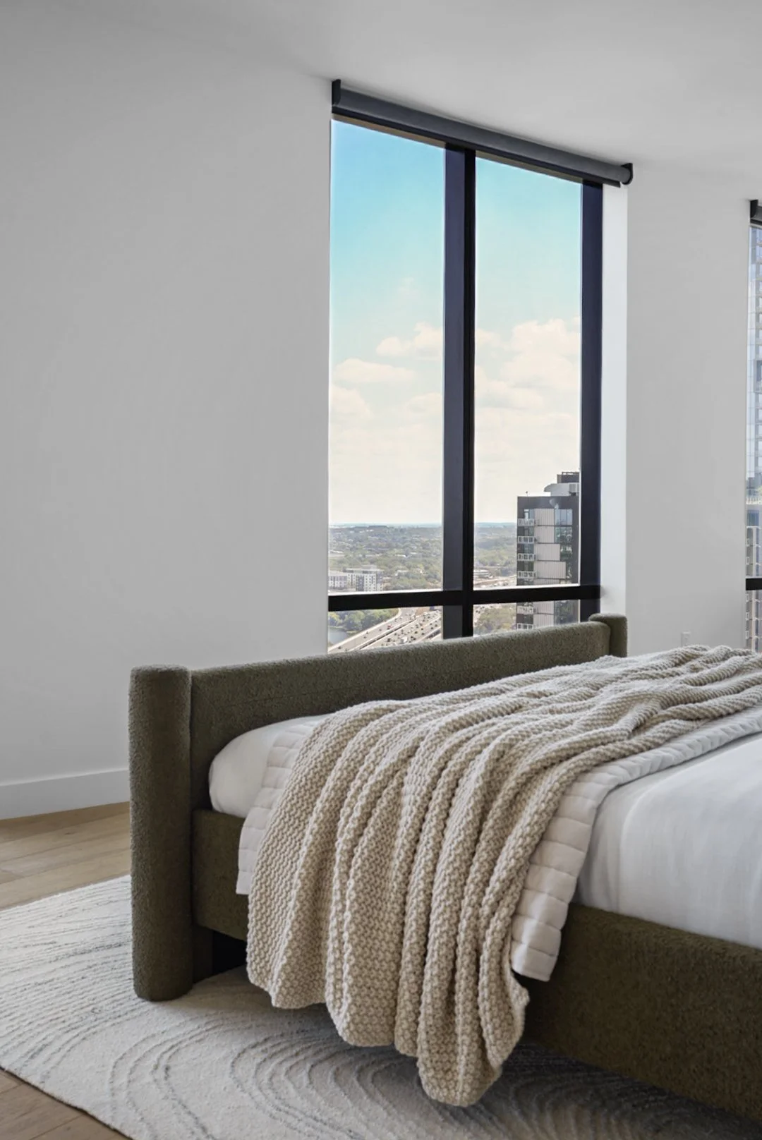 Modern bedroom with a large window showing a city view, a bed with a beige upholstered headboard, white bedding, and a chunky knitted blanket, on a textured rug.