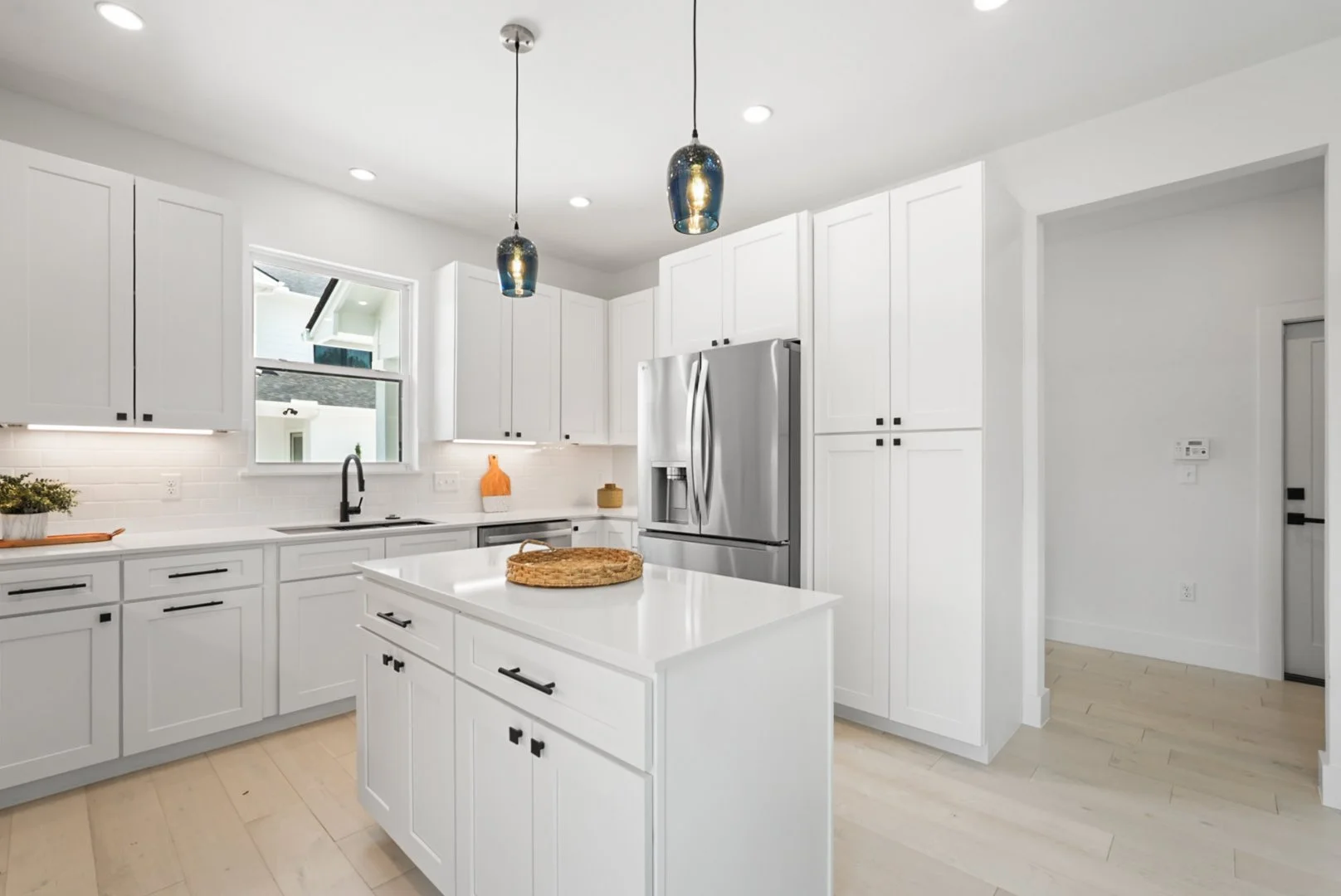 Modern white kitchen with pendant lights, stainless steel refrigerator, white cabinets, black hardware, and light wood flooring.