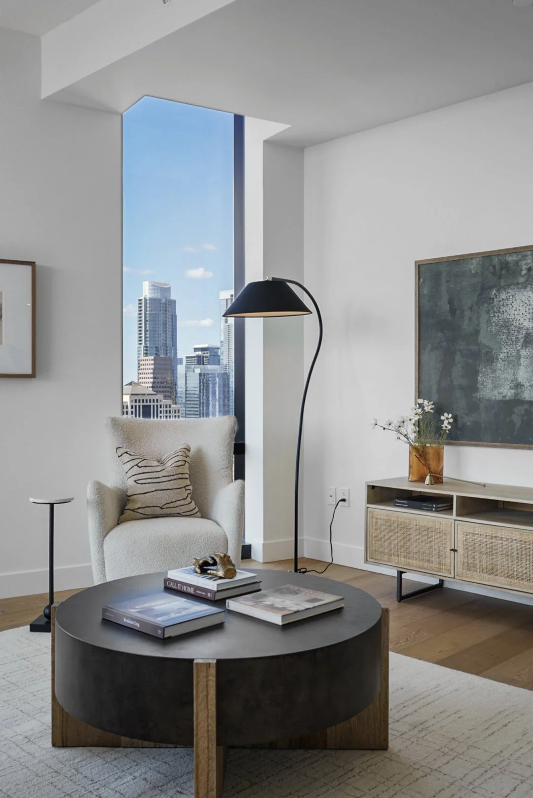 Modern apartment living room with a large window showing city skyscrapers, white armchair with striped cushion, black arc floor lamp, wooden media console with a potted plant, and round coffee table with books and decorative items.