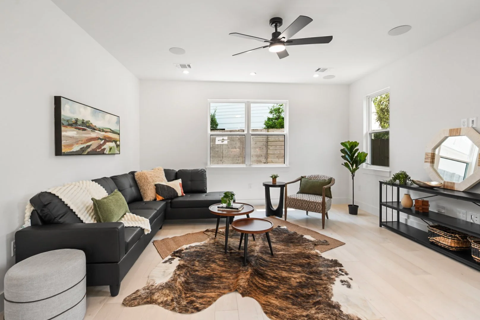 Modern living room with a black leather sectional sofa, a cowhide rug, a small round coffee table, and multiple armchairs. The room features white walls, light hardwood floors, a ceiling fan, and large windows with views of a wooden fence and greener