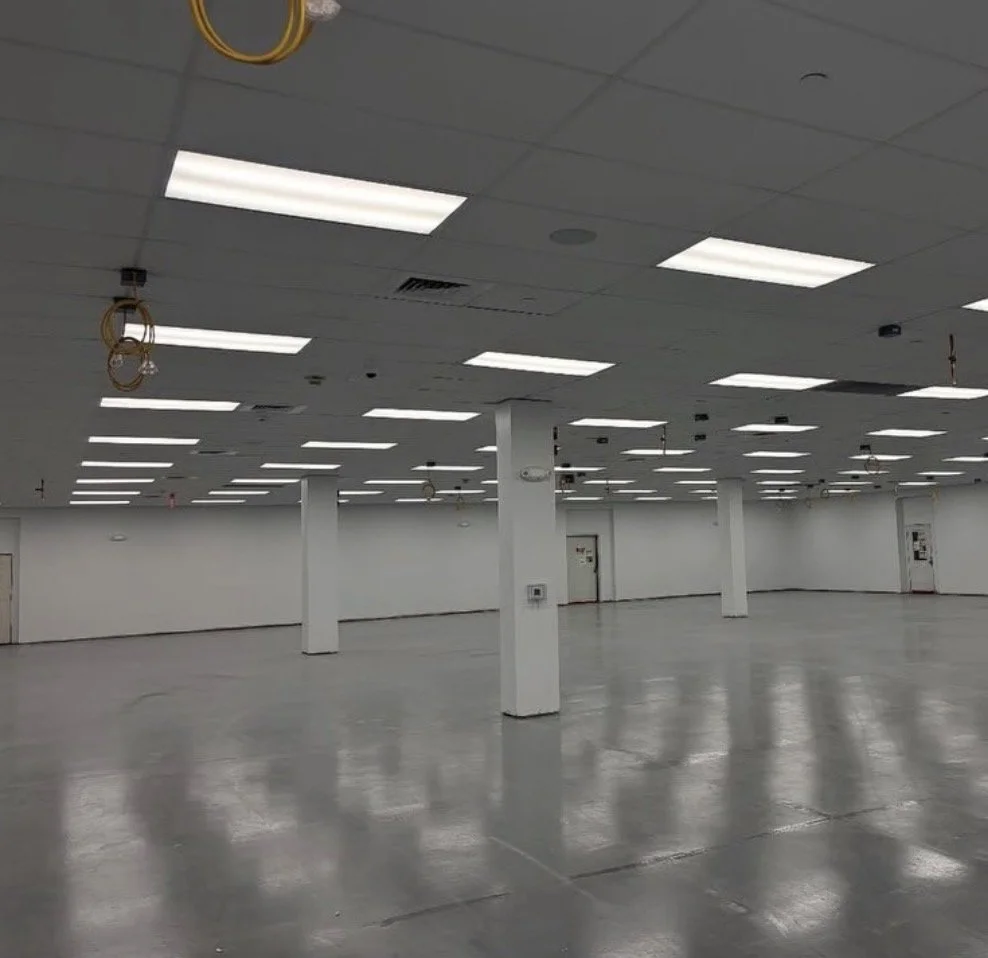 Empty commercial or industrial space with white walls, gray concrete floor, ceiling lights, and support columns.