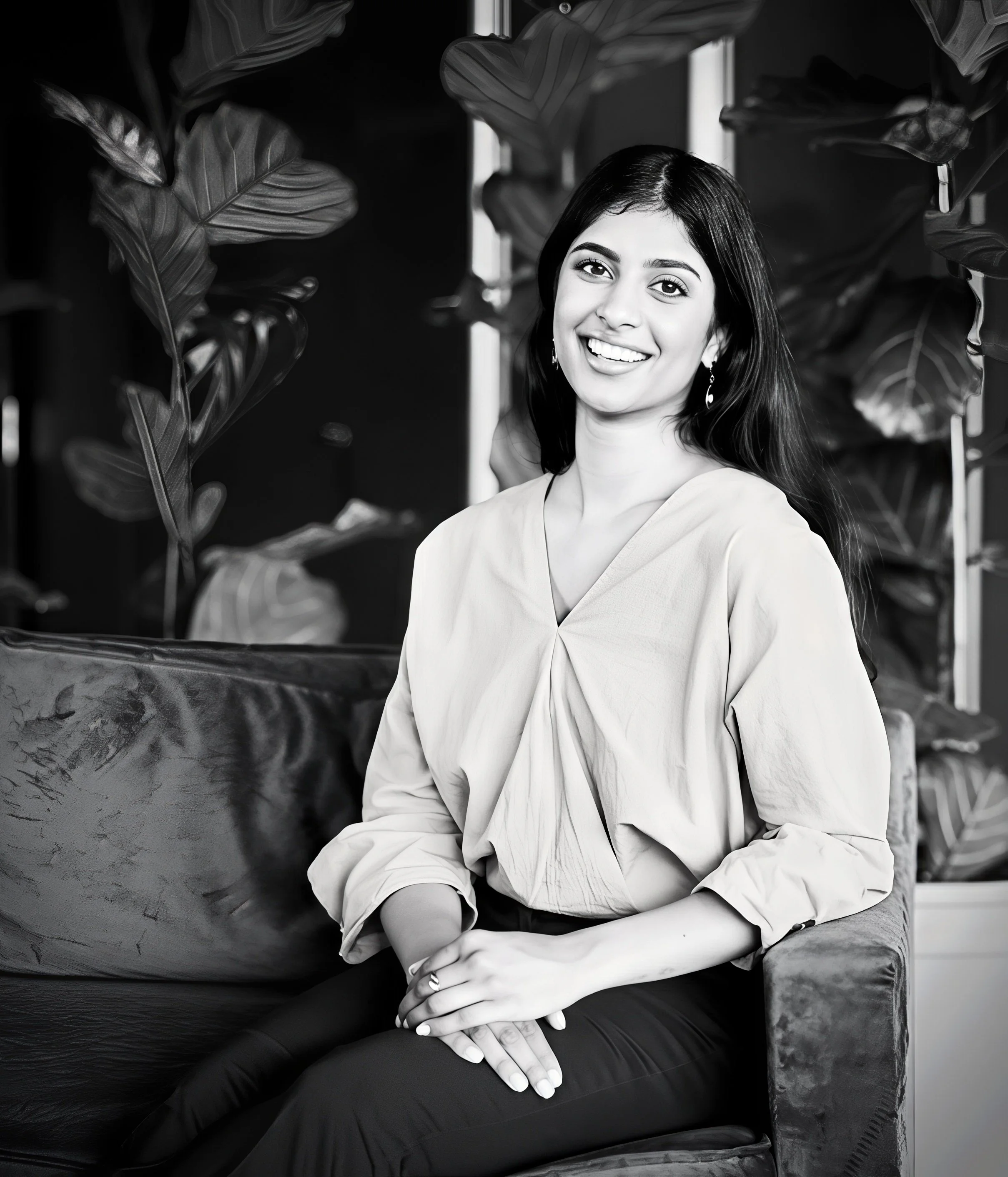 A smiling woman with dark hair sitting on a sofa in front of large leafy plants, in a black and white photo.