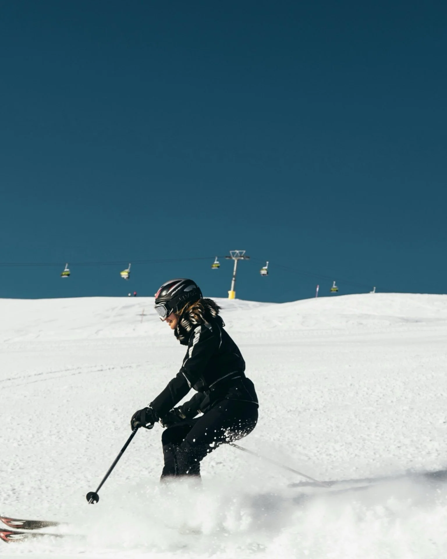 Person skiing on snow-covered slope with ski lift in the background under clear blue sky.