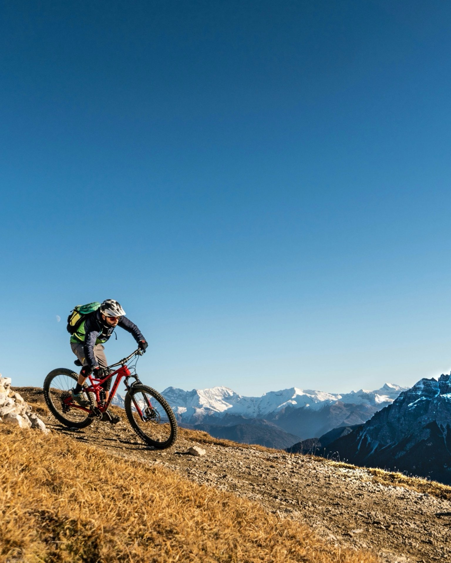 A mountain biker riding downhill on a dirt trail with snow-capped mountains in the background under a clear blue sky.
