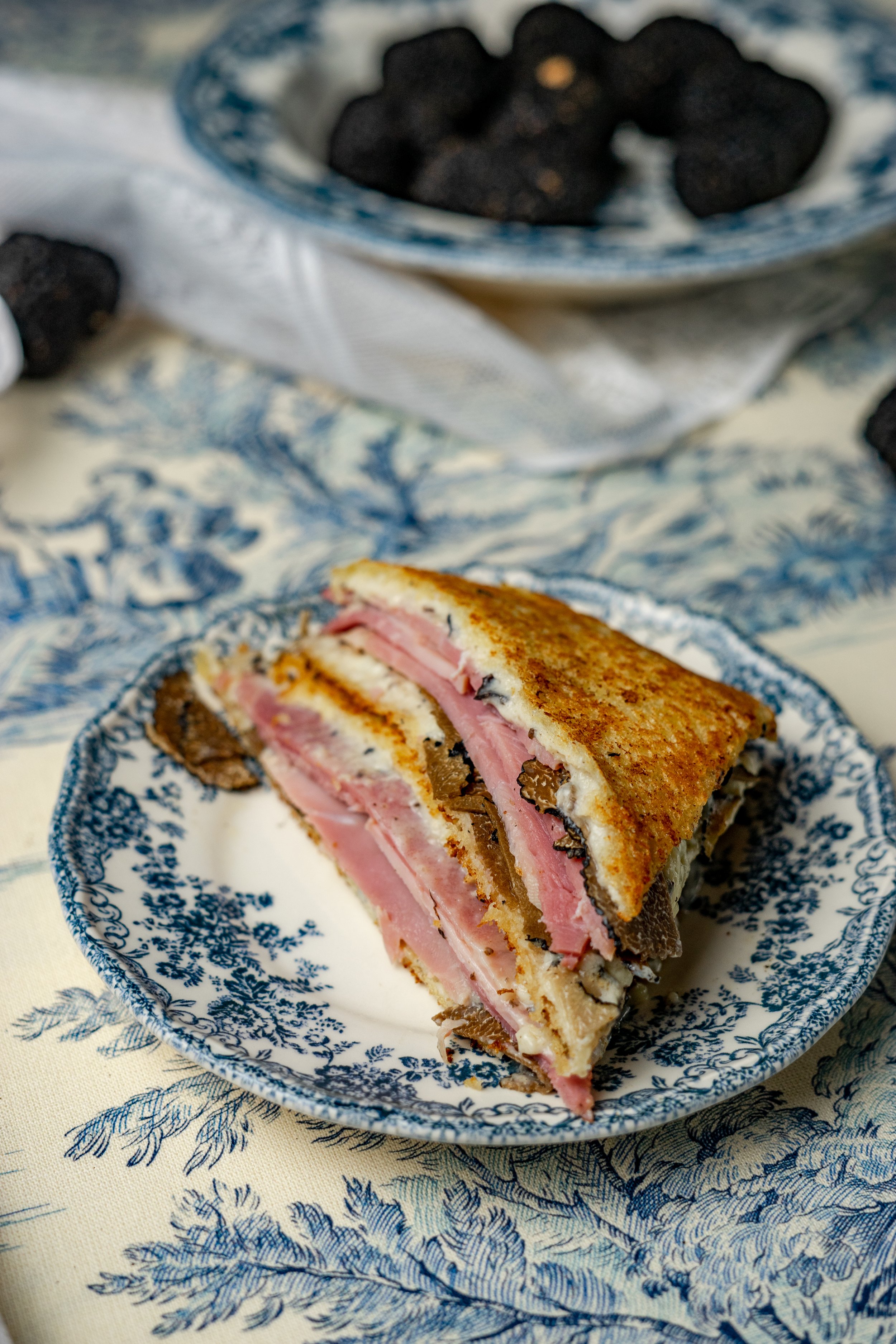 Sandwich avec jambon, champignons et fromage sur une assiette en porcelaine à décor bleu, posé sur une nappe ivoire avec un motif floral bleu, avec un bol contenant des truffes en arrière-plan.