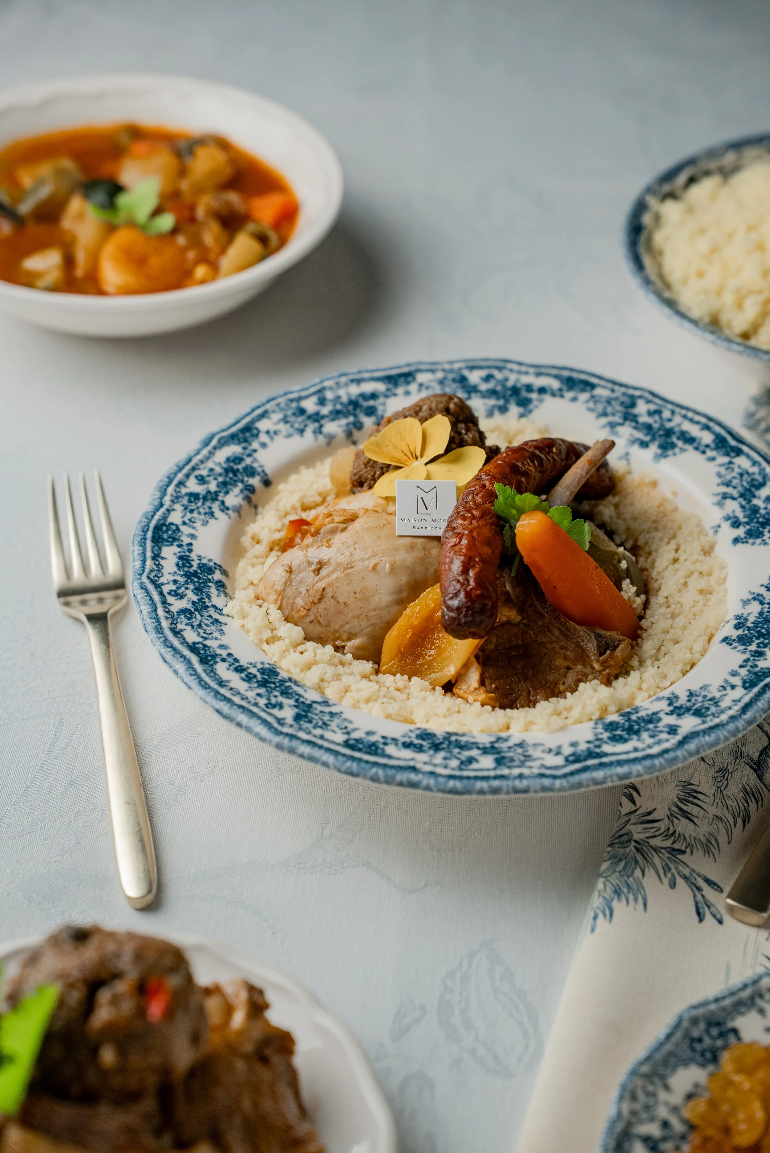 Un plat traditionnel avec couscous, légumes, viande et saucisse, décoré d'une fleur en garniture, entouré de plusieurs autres plats et couverts.