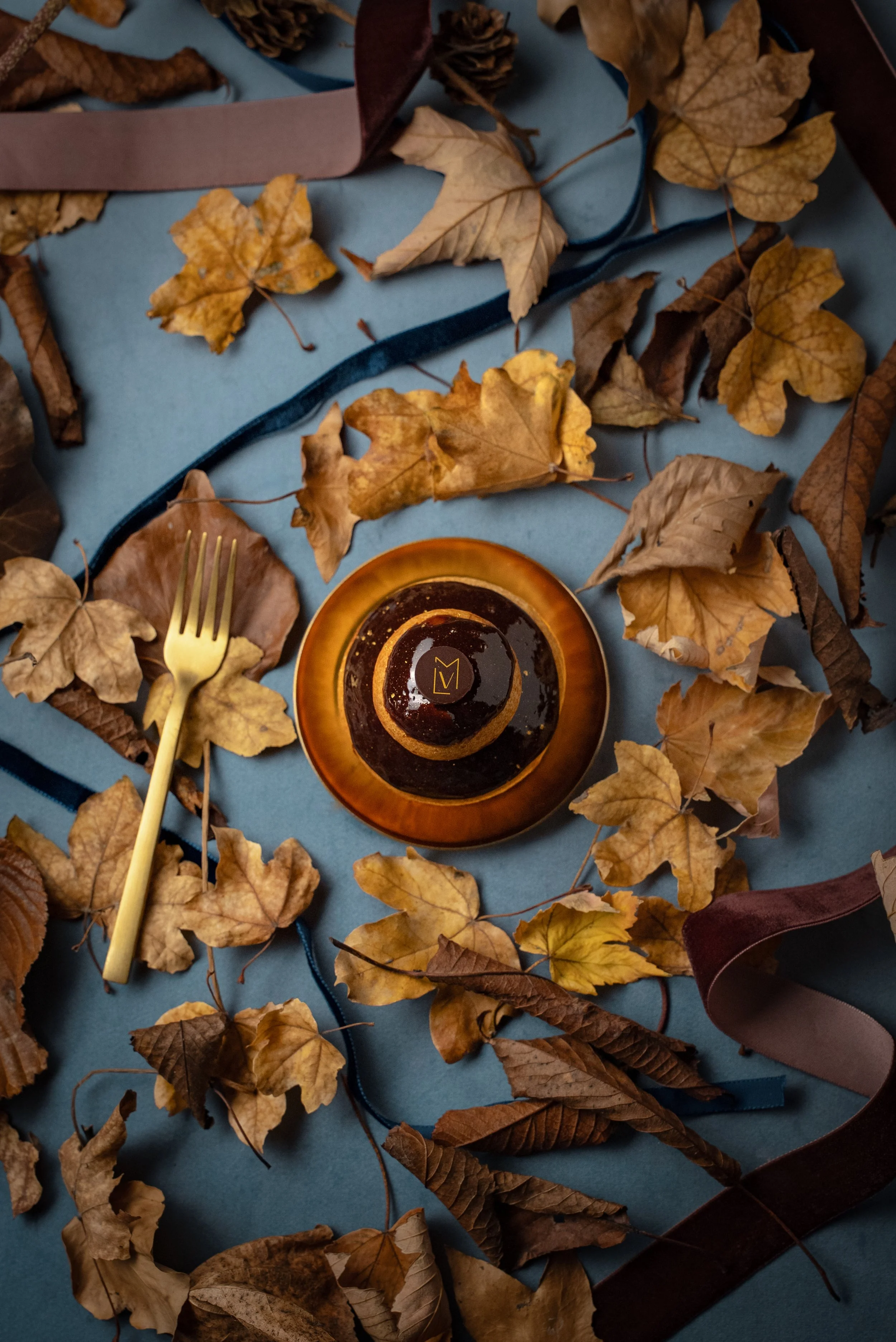 Une photographie d'automne avec des feuilles sèches de différentes couleurs dispersées sur une surface bleue. Au centre, une petite assiette orange contenant un dessert glacé décoré avec un cercle de caramel et un logo en or 'LV'. Sur le côté, une fourchette dorée repose parmi les feuilles.