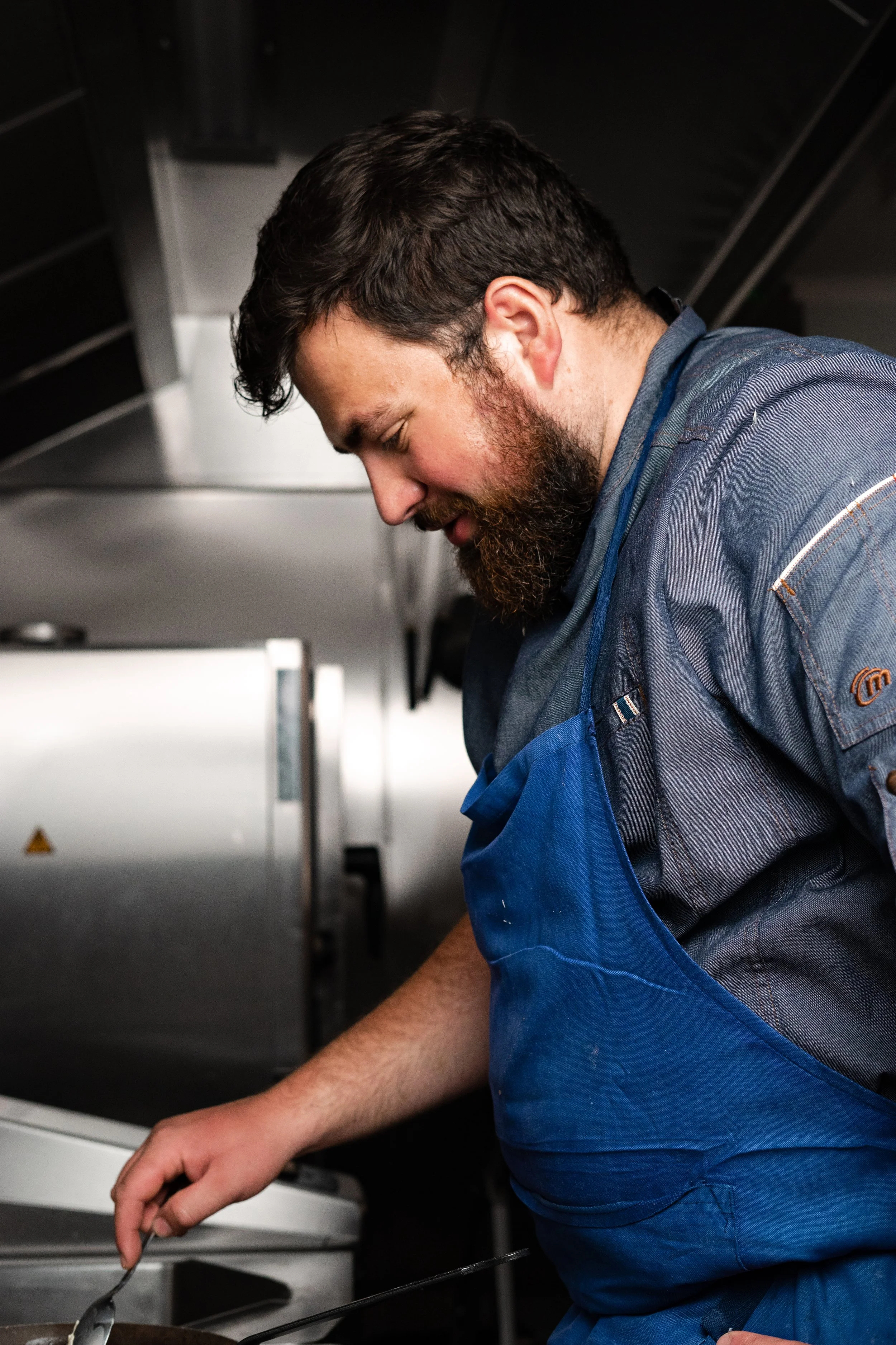 Un homme coiffé de cheveux foncés et une barbe, portant un uniforme de chef cuisinier avec une bavette bleue, en train de cuisiner dans une cuisine professionnelle