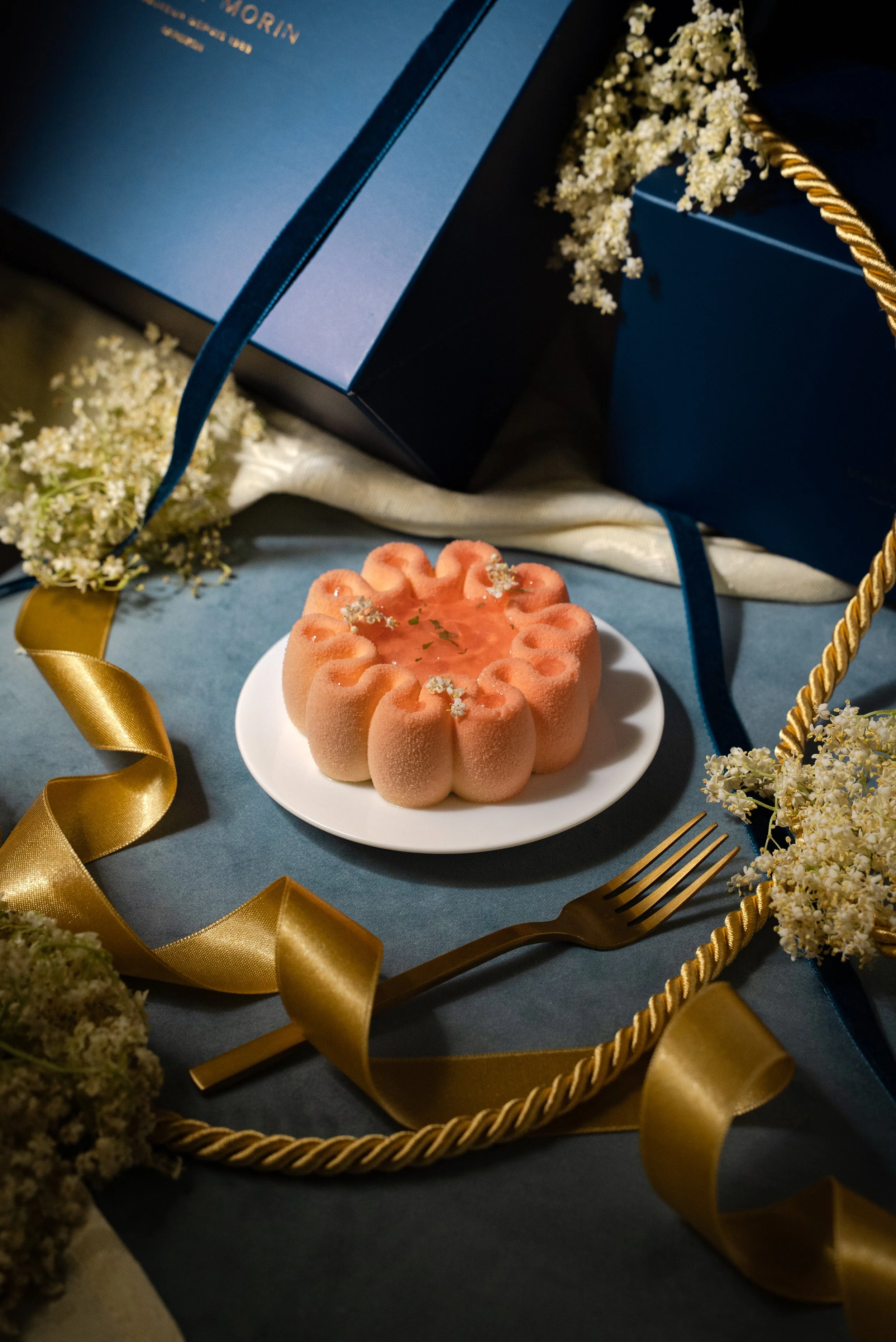 Un gâteau en forme de fleur sur une assiette blanche, entouré de décorations festives dorées et bleues, avec des fleurs blanches et un fourchette dorée.