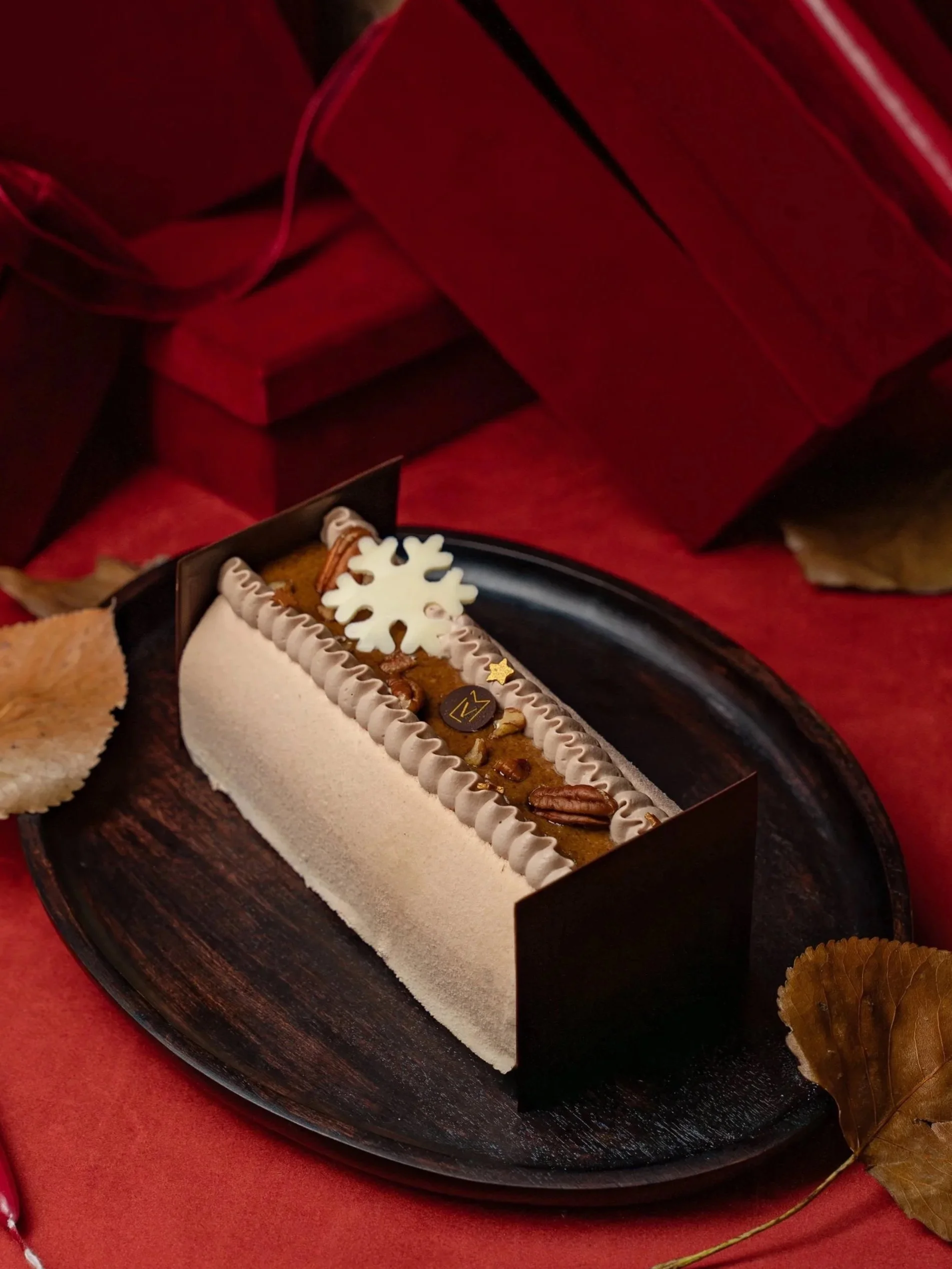 Gâteau de Noël décoré avec un flocon de neige en chocolat blanc, des noix et une garniture caramel, présenté sur une assiette noire avec des feuilles d'automne en décor.