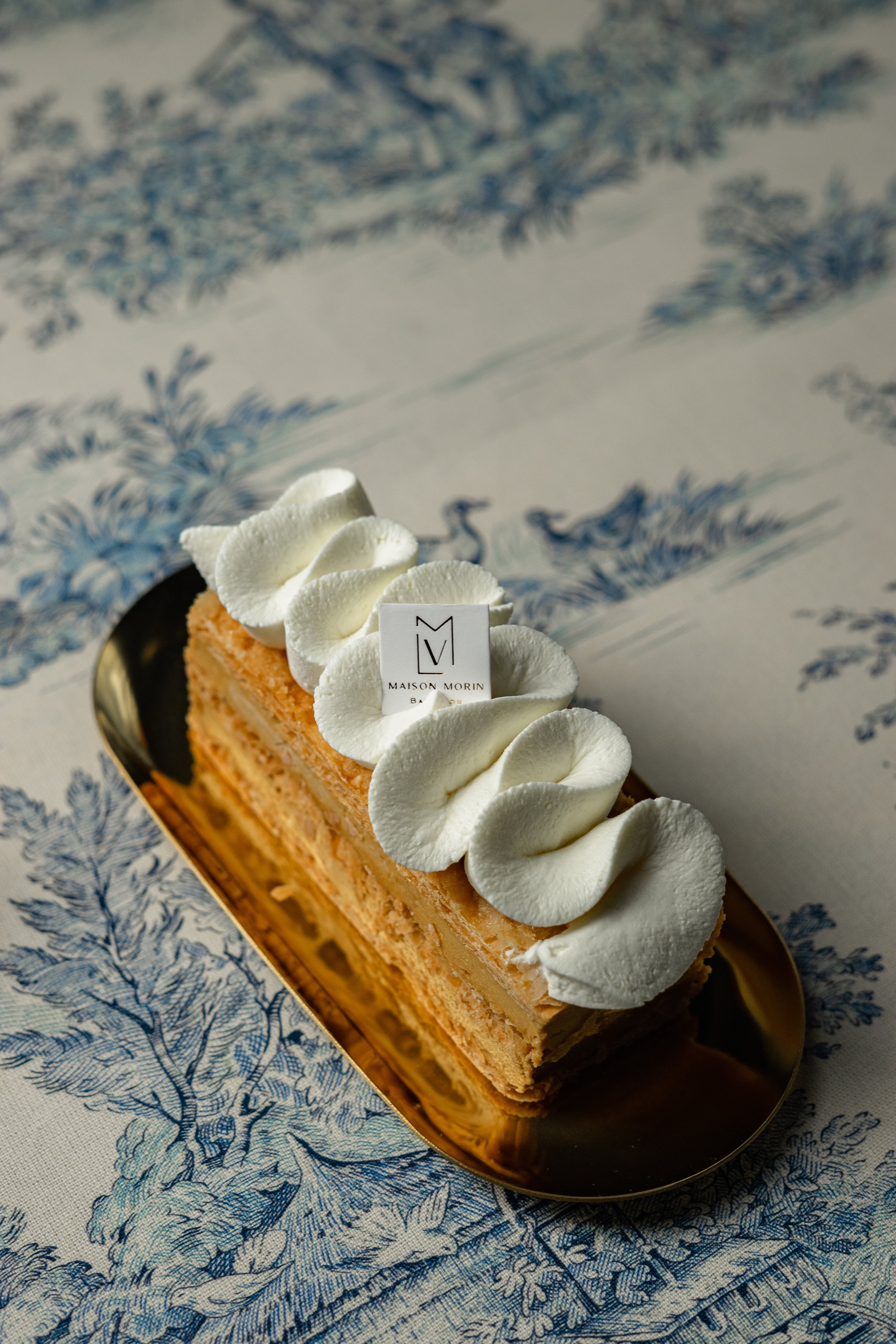 Gâteau pâtisserie rectangular avec des couches de biscuit et de crème, décoré de crème fouettée enroulée en haut, avec un petit drapeau en papier portant le logo 'Maison Morin'.