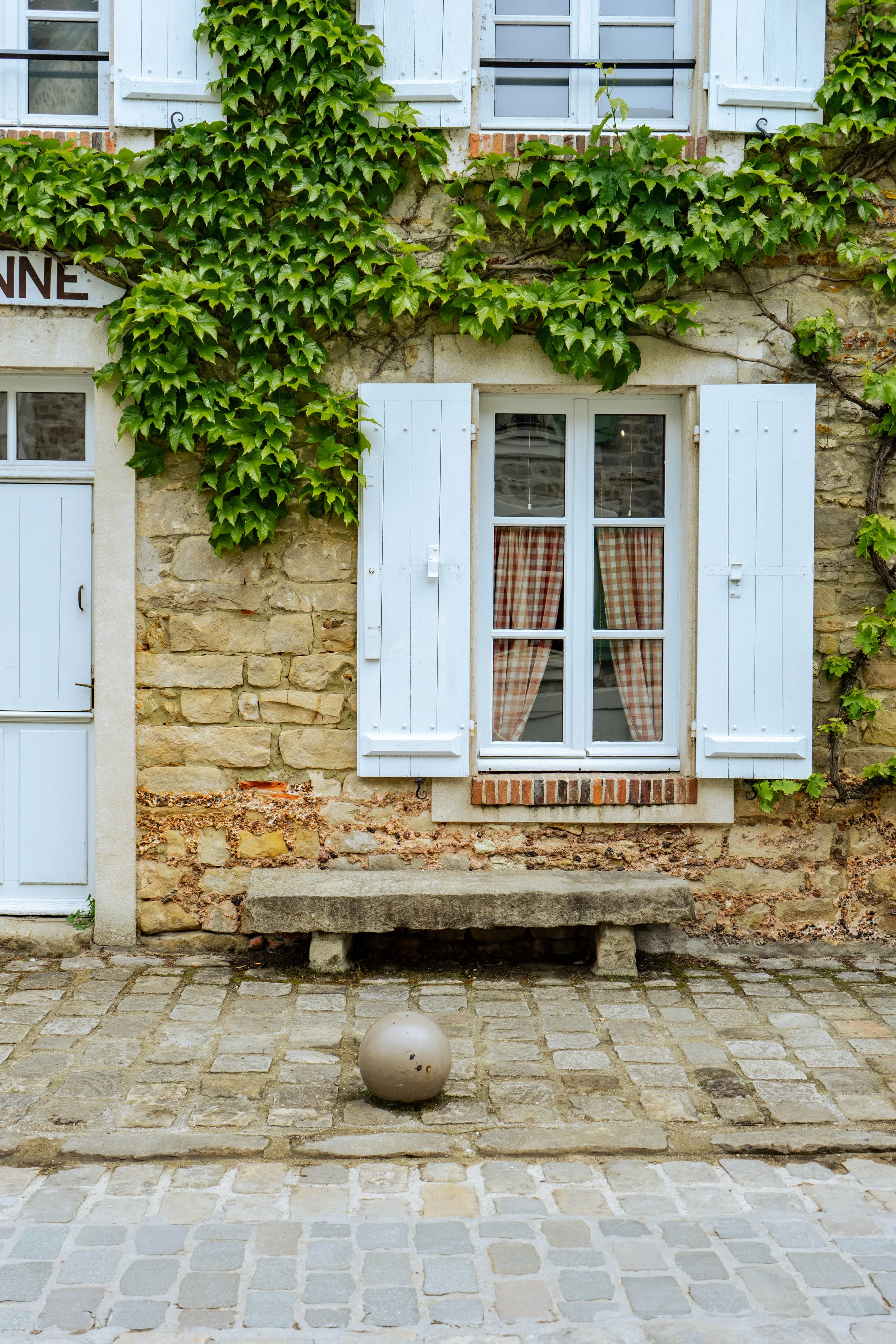 Fenêtre avec volets blancs, rideaux à carreaux, maison en pierre avec des volets en bois peints en blanc, plantes grimpantes vertes, pavés en pierre et un banc en pierre en bas.