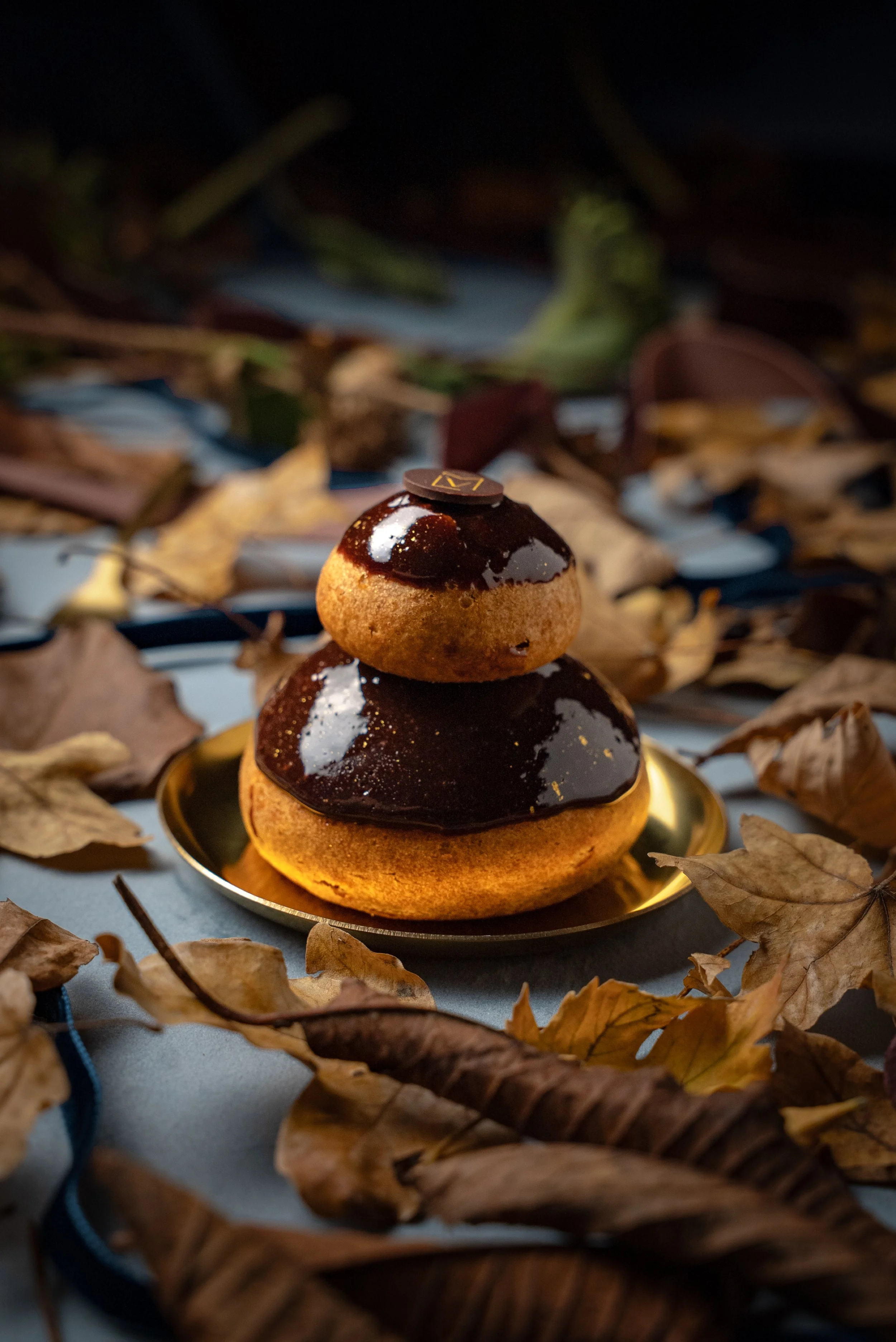 Gâteau en forme de pierre avec deux niveaux recouverts de glaçage au chocolat, posé sur une petite assiette dorée, entouré de feuilles mortes.