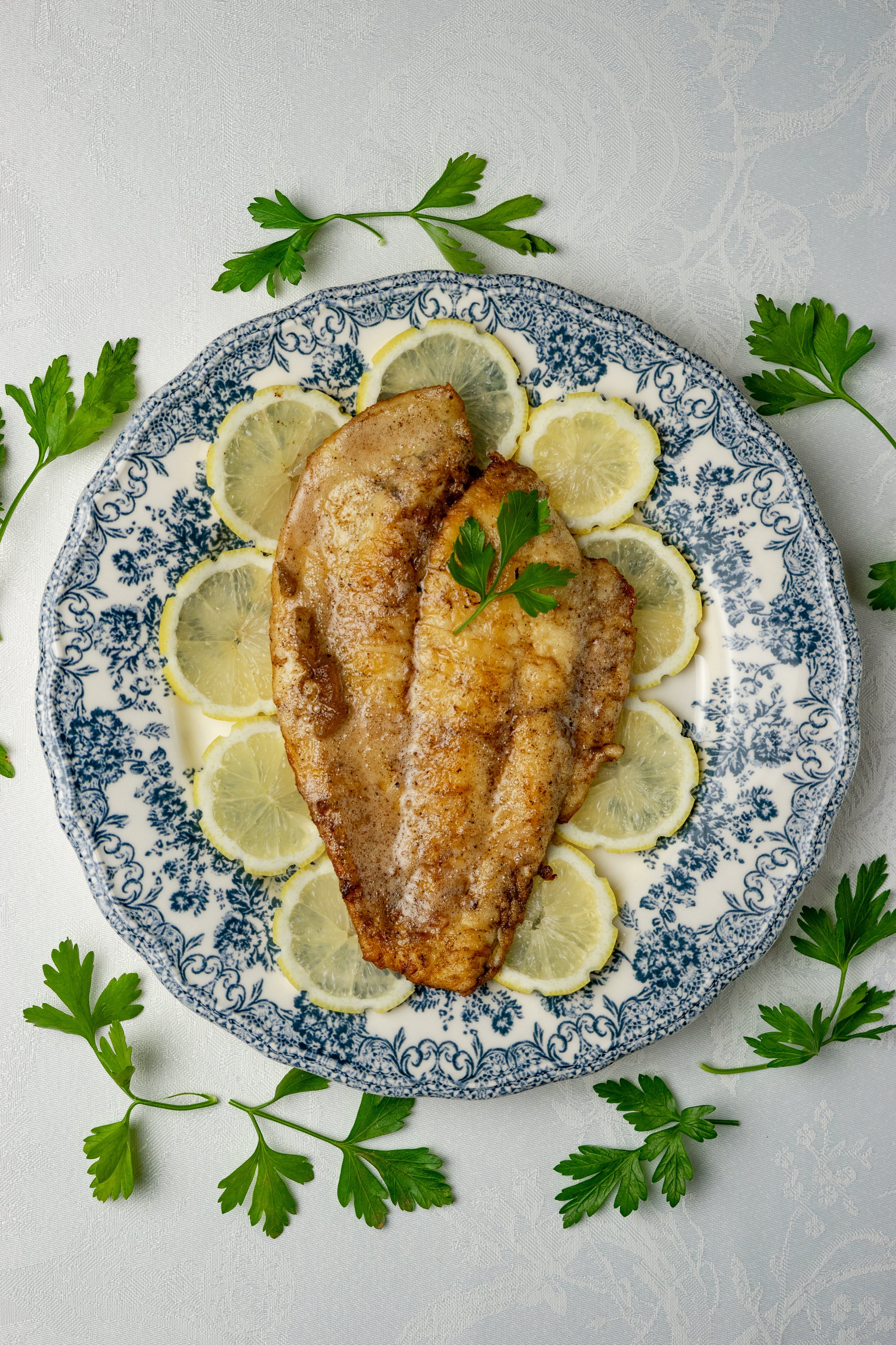 Filet de poisson frit avec rondelles de citron et herbes fraîches sur une assiette en porcelaine bleue, entouré de feuilles de persil.