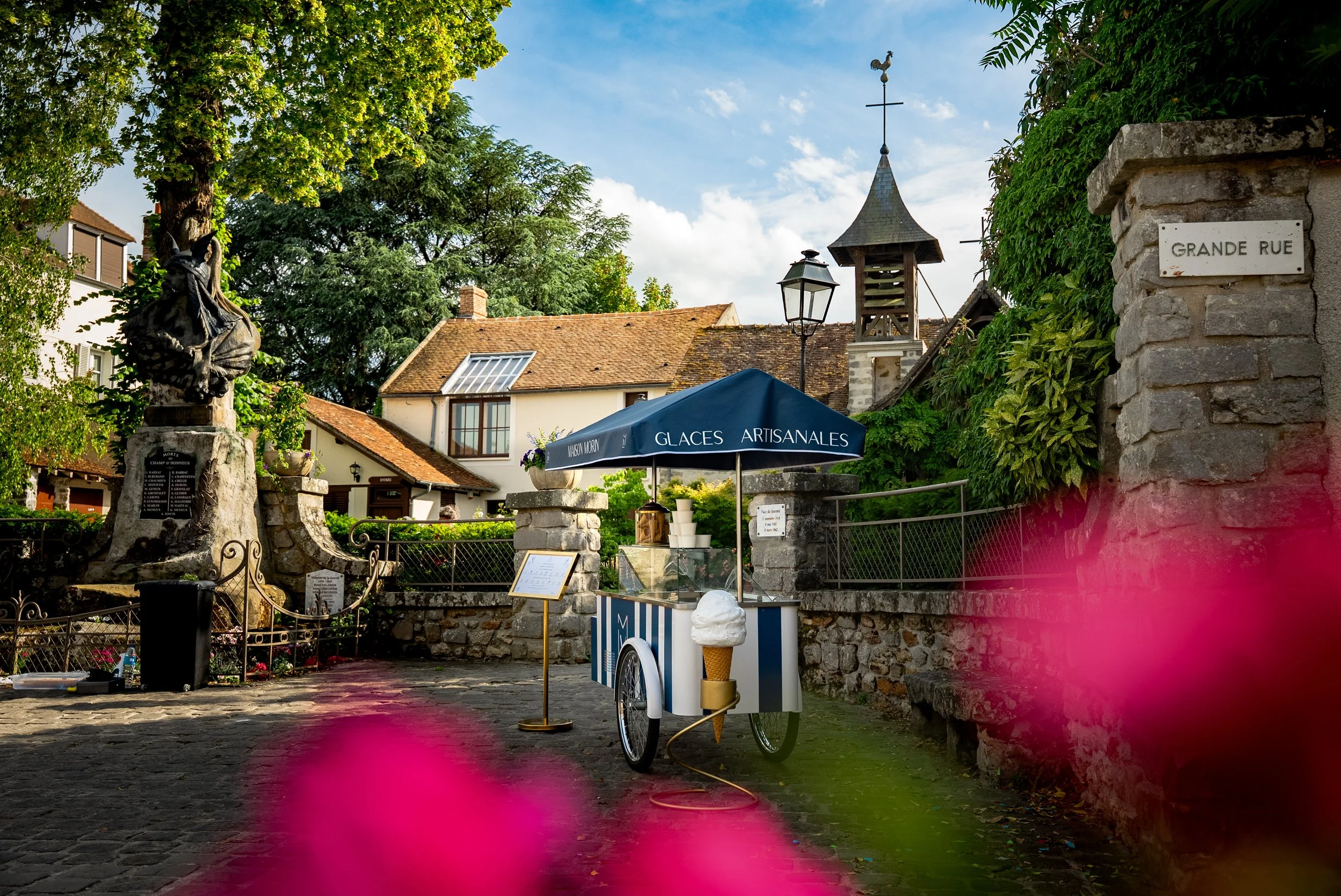 Un stand ambulant de glace artisanale avec une large ombrelle bleue portant l'inscription "Glaces Artisanales" dans une rue pavée. En arrière-plan, il y a une statue en pierre d'un homme avec une plaque sombre, un lampadaire, une petite église en pierre avec un clocher, et des maisons avec des toits en tuiles. Le tout est entouré de murs en pierre et de végétation dense.