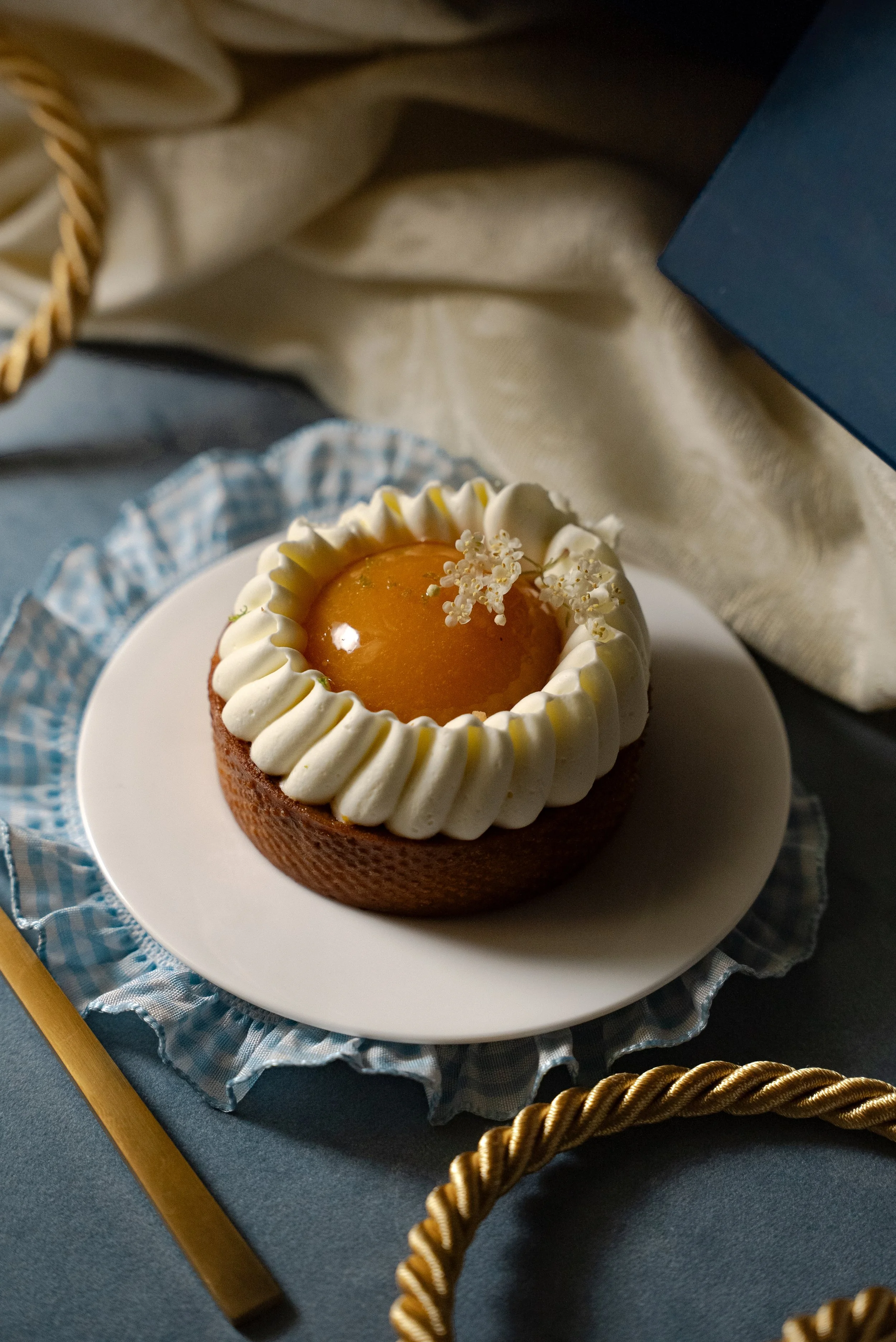 Gâteau décoré avec une cerise en gelée au centre, entourée de crème fouettée et de petites fleurs blanches, posé sur une assiette blanche avec un fond de tissu beige et d'autres éléments décoratifs.