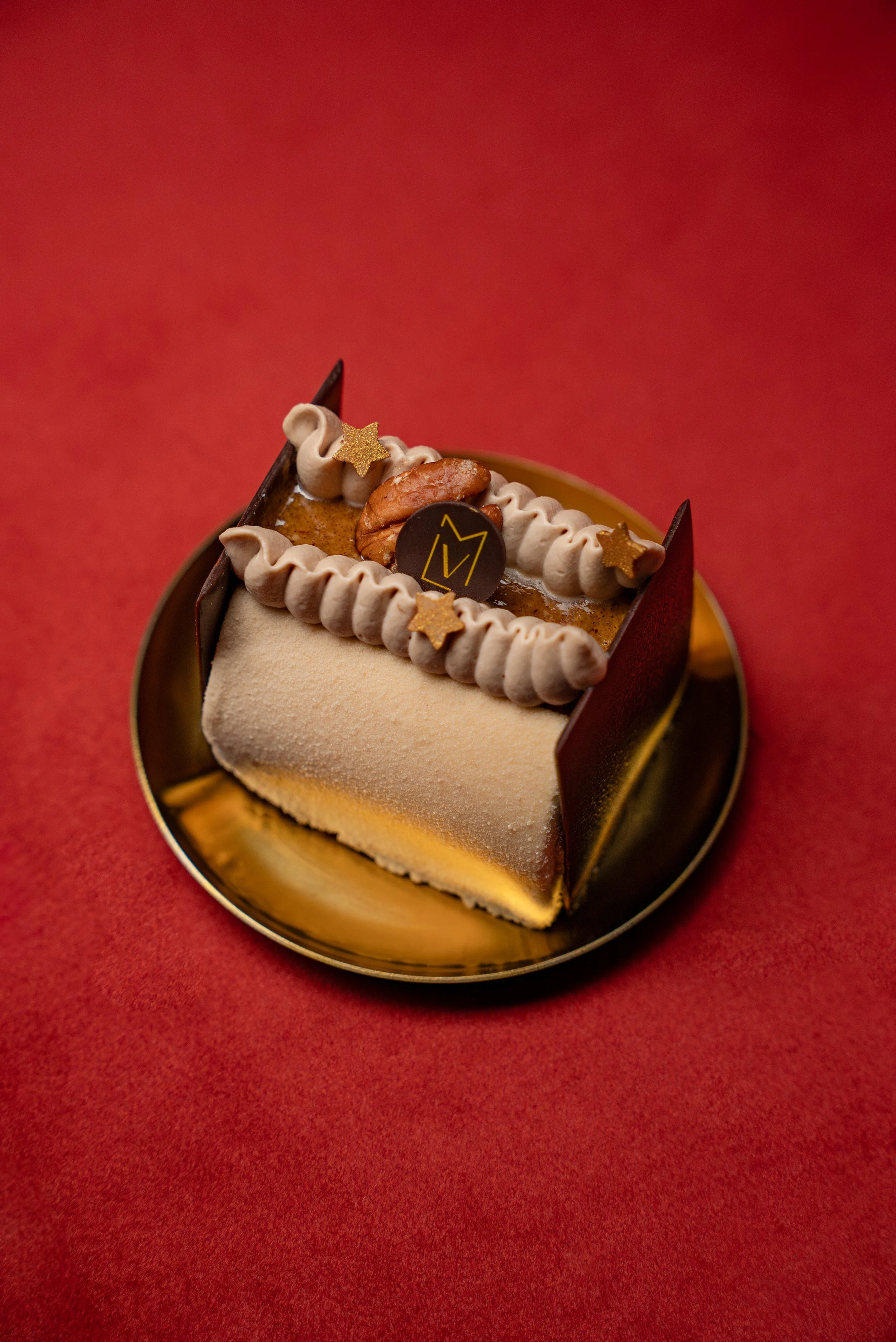 Gâteau décoré avec des étoiles dorées, un fruit sec, une pièce en chocolat avec un logo et une crème en forme de ruban, dans un plateau doré sur fond rouge.