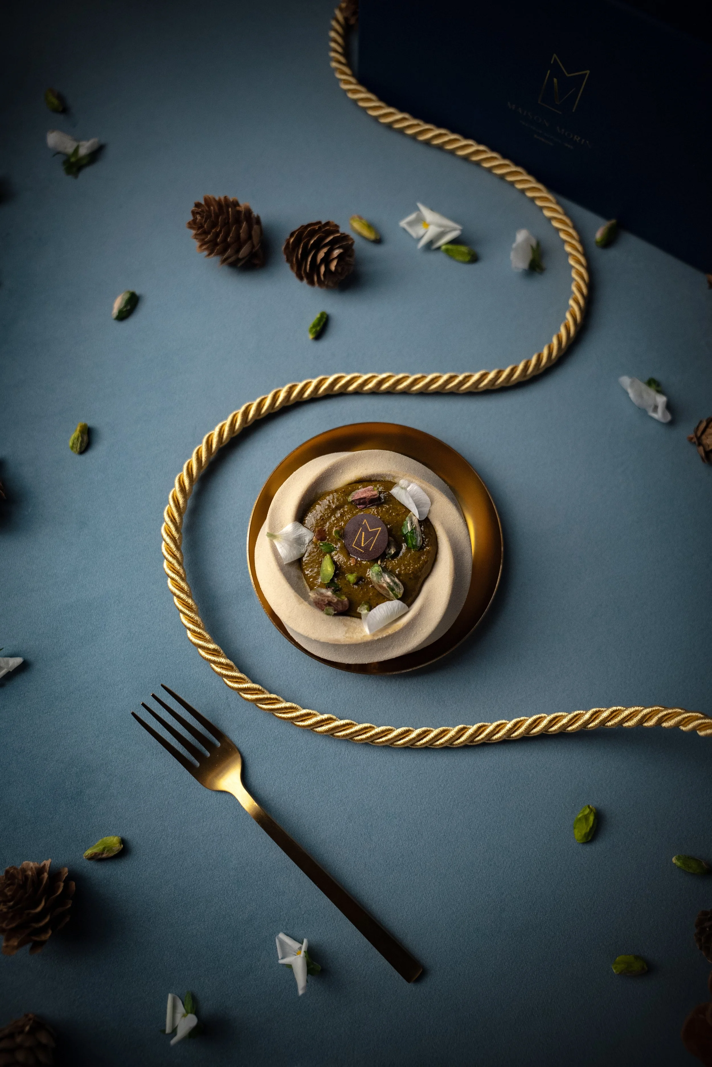Dessert élégant avec un motif de pistache, décoré de pétales de fleurs blanches, présenté sur une assiette dorée, entouré de cordes dorées, avec des pommes de pin et des pétales de fleurs blanches en décoration sur une surface bleue.