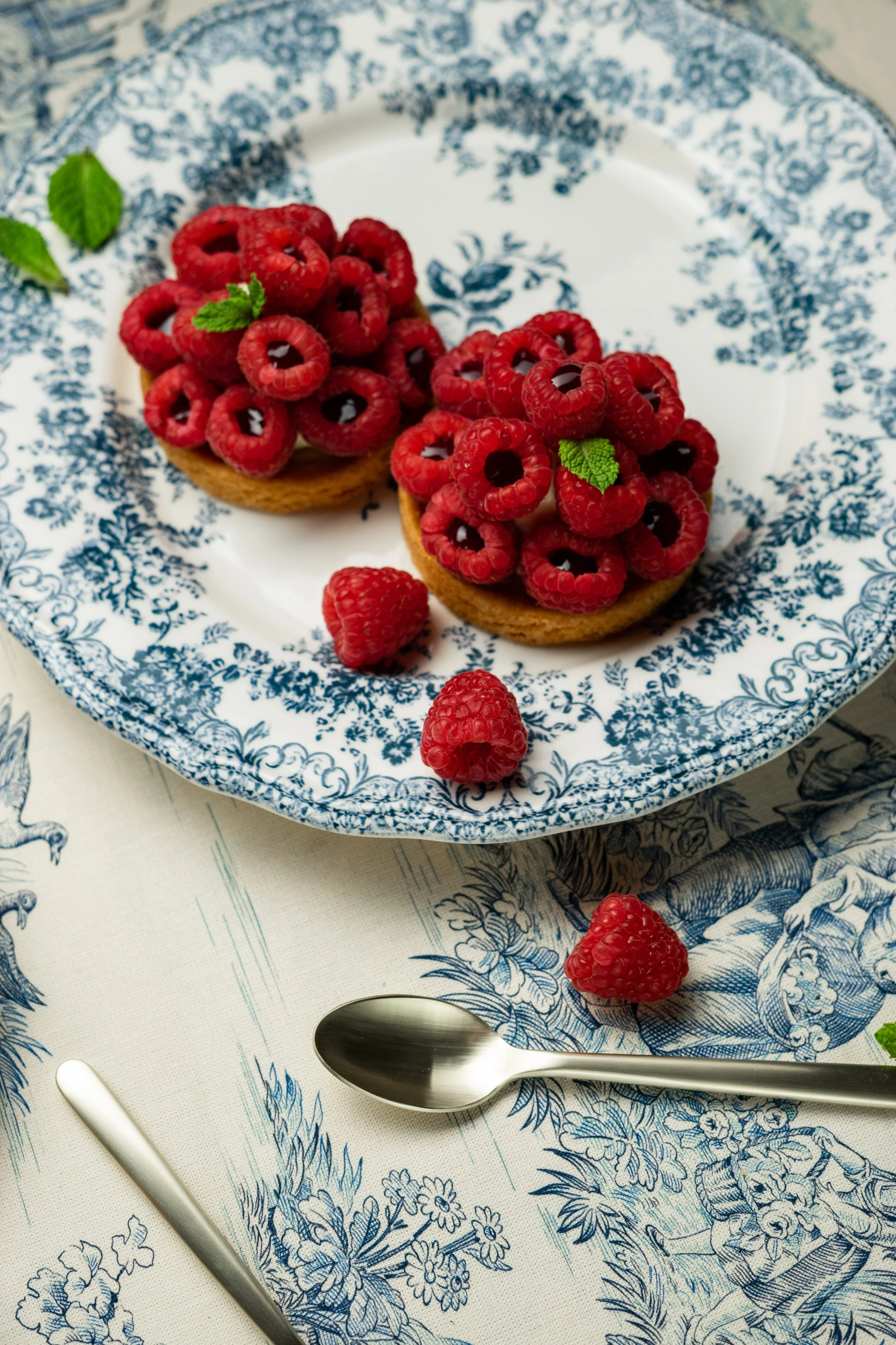Deux tartines de biscuits beurrés garnies de framboises fraîches avec des feuilles de menthe, sur une assiette à motifs bleus et blancs; quelques framboises et une cuillère en métal sont aussi sur la nappe avec un motif floral bleu.