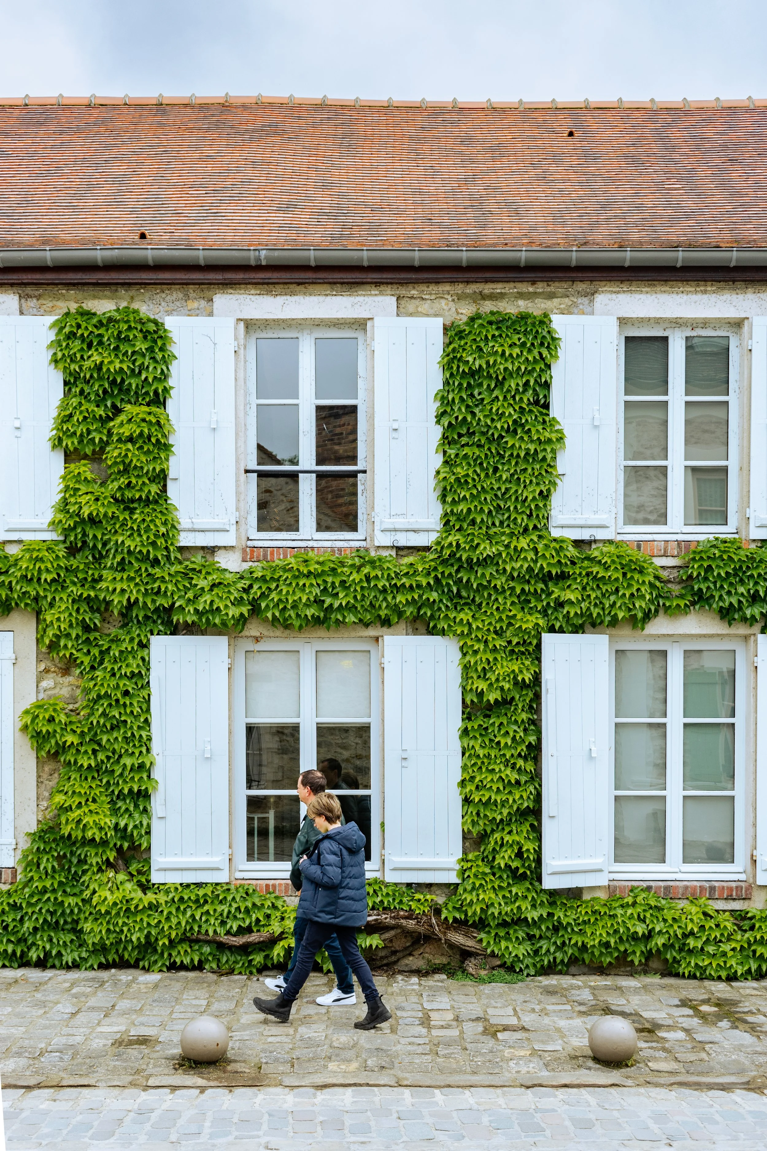 Deux personnes marchant devant un bâtiment avec des volets en bois blanc et une façade recouverte de lierre vert.