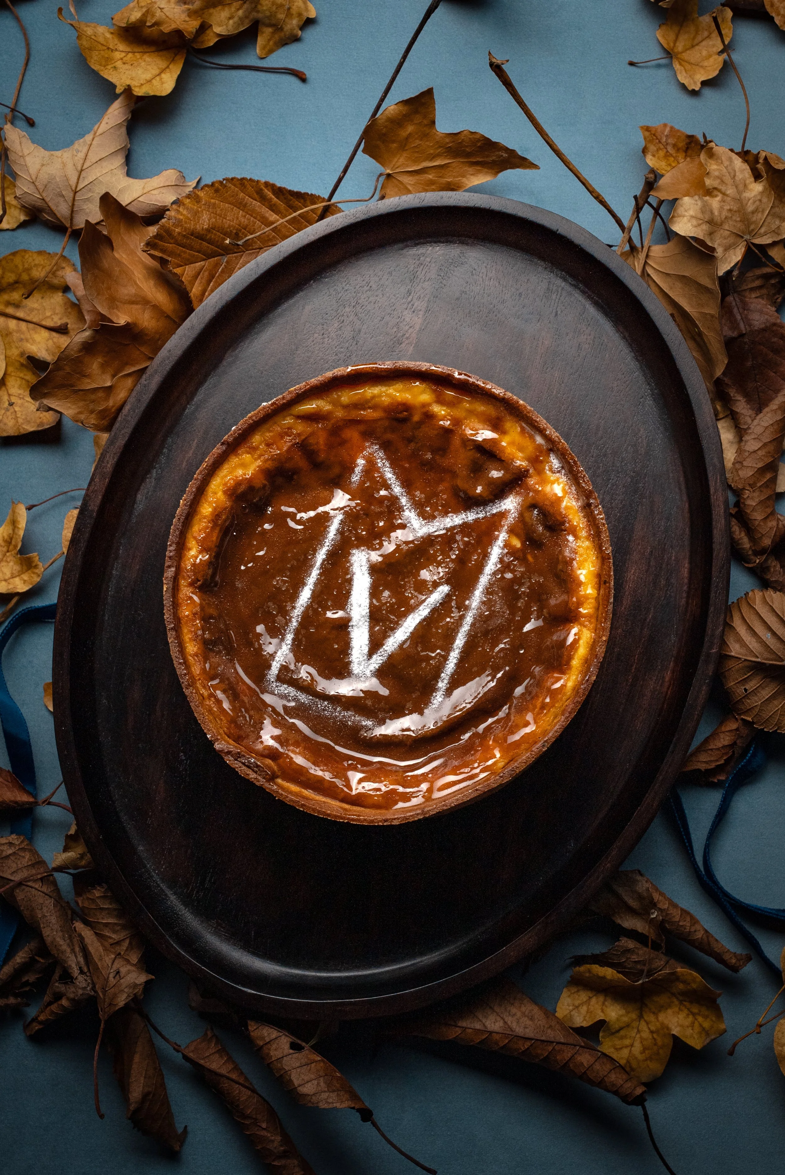 Gâteau aux pommes avec des symboles dessinés à la craie blanche dessus, entouré de feuilles mortes en bois et sur un fond bleu.