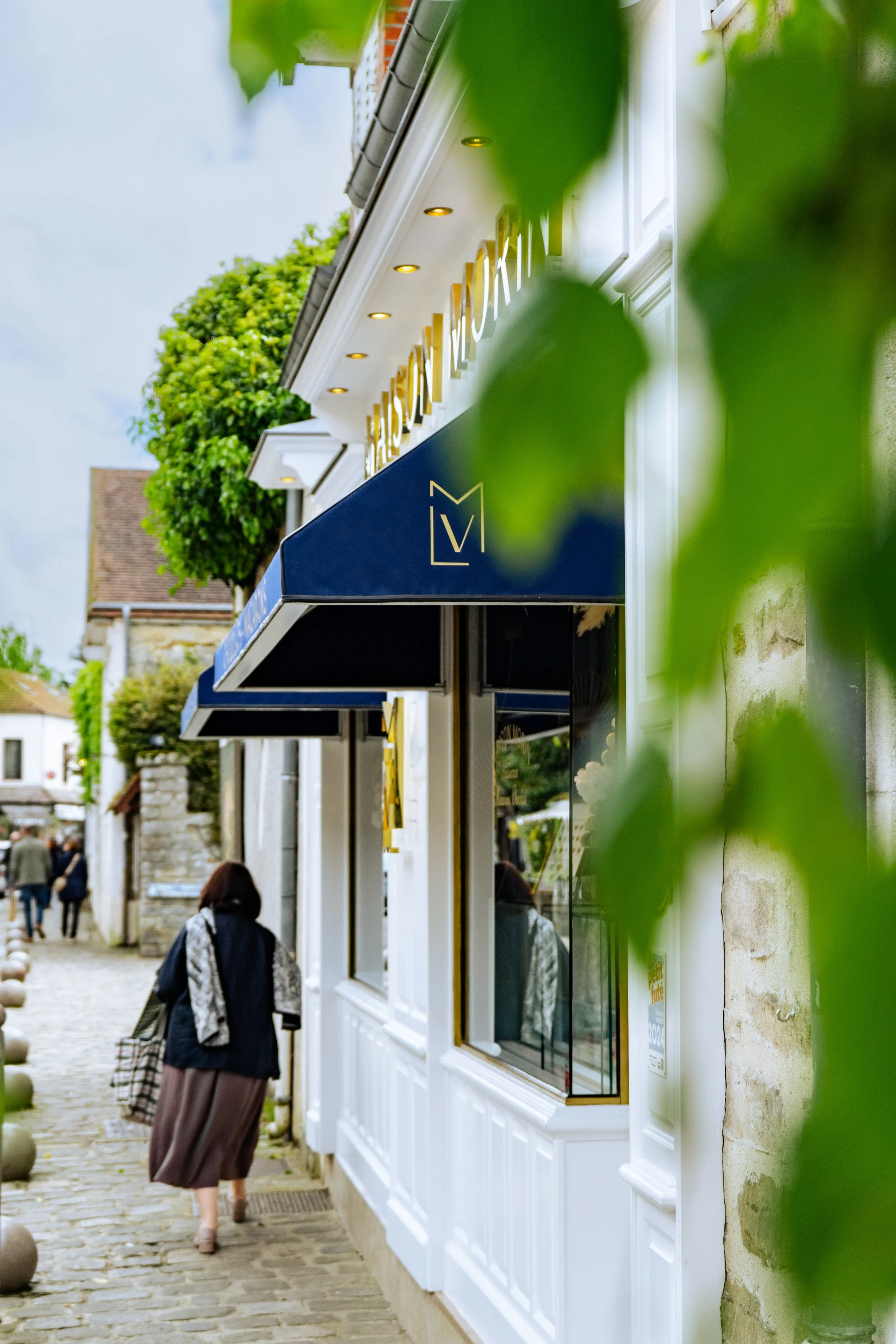 Une femme marche dans une rue pavée près d'une boutique avec une enseigne bleue et une vitrine, entourée de verdure, en journée.